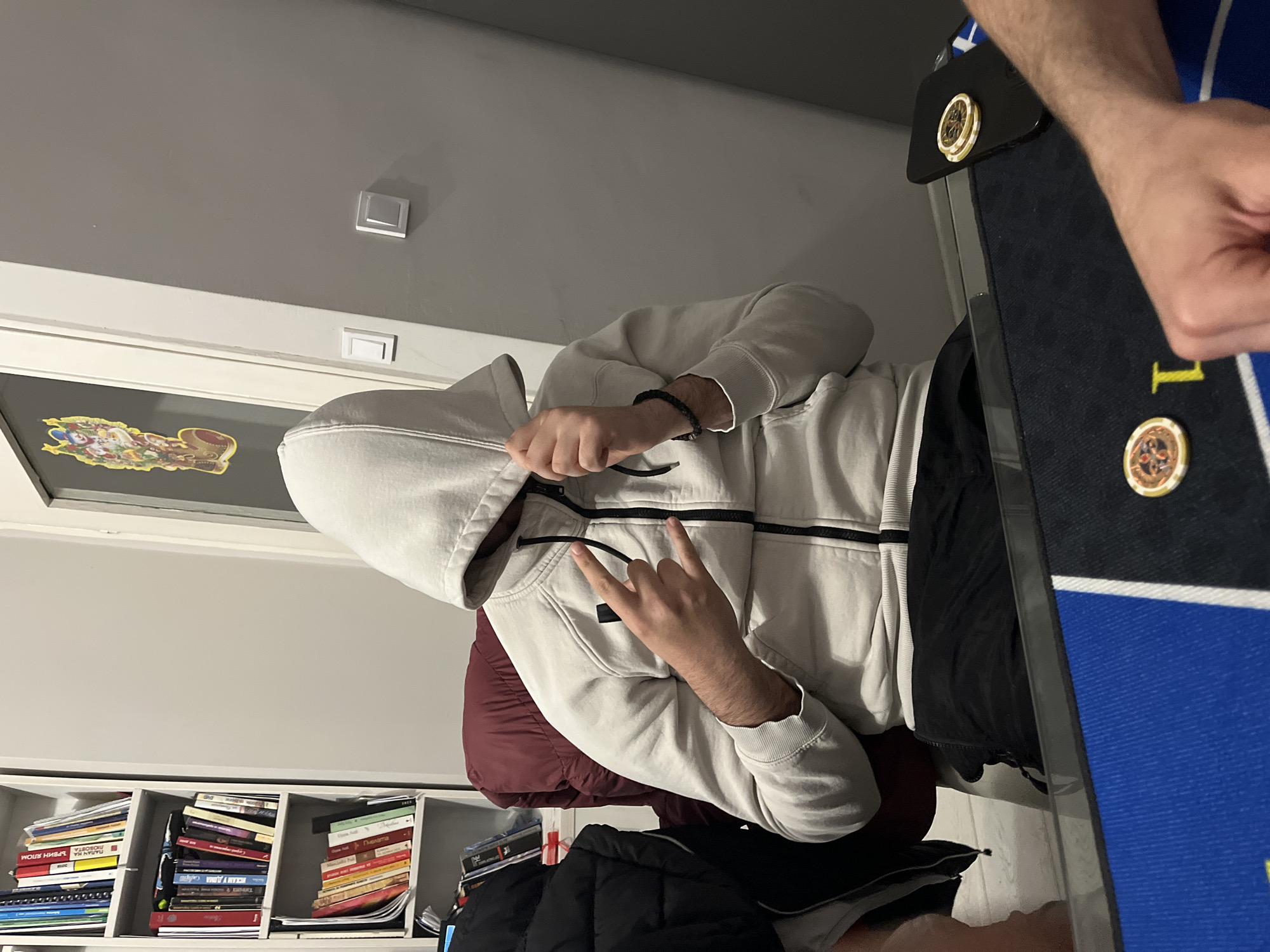 The photograph depicts a person seated in a chair, wearing a light-colored hooded sweatshirt. The individual is making a hand gesture, possibly a peace sign or rock-on hand gesture. A poker table is present in the frame. The environment includes a bookshelf filled with books, and a window with a decorative sticker. Based on the architectural elements visible such as wall mounted lights, the location is likely indoors. The details provided do not allow to identify this exact location in Burgas, Bulgaria.