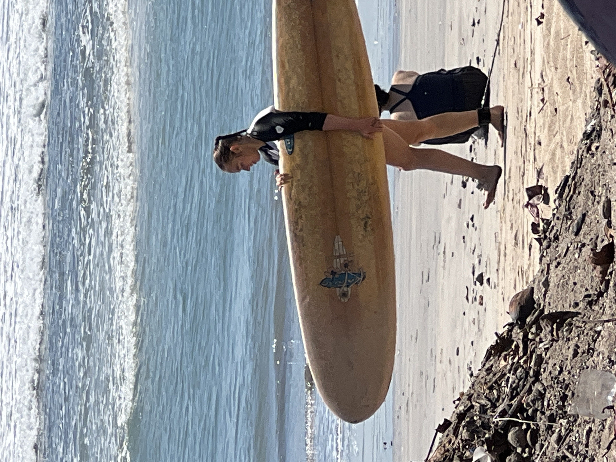 A woman walks on the beach in Kuta, Indonesia, carrying a surfboard. She is wearing a black and white wetsuit and her hair is pulled back in a ponytail. The surfboard is a light tan color with a blue and white logo on it. In the background, the ocean is visible with waves crashing on the shore. Another person is sitting on the sand in the background, facing away from the camera. In the foreground, there is a pile of debris on the beach, including rocks, sticks, and plastic. The sand is light brown and there are a few small rocks scattered across it. The sky is bright and sunny.
