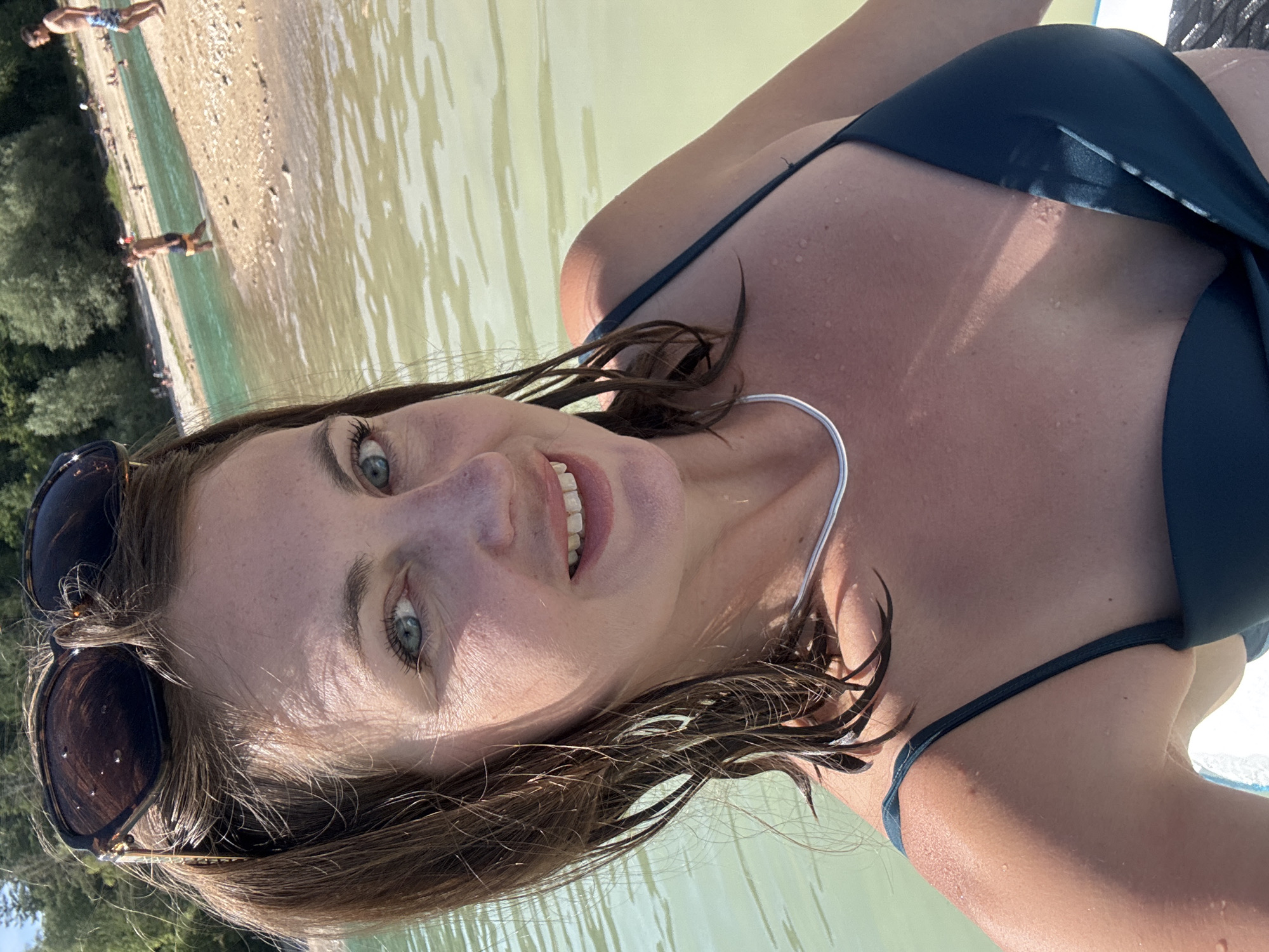 A woman with brown hair and blue eyes smiles at the camera. She is wearing a dark green bikini and sunglasses pushed up on her head. The woman is in the water, which appears to be a lake or river. In the background, there are trees and a sandy beach with several people. The location is the Lac de Vouglans in Maisod, France. The lighting suggests it is a sunny day.
