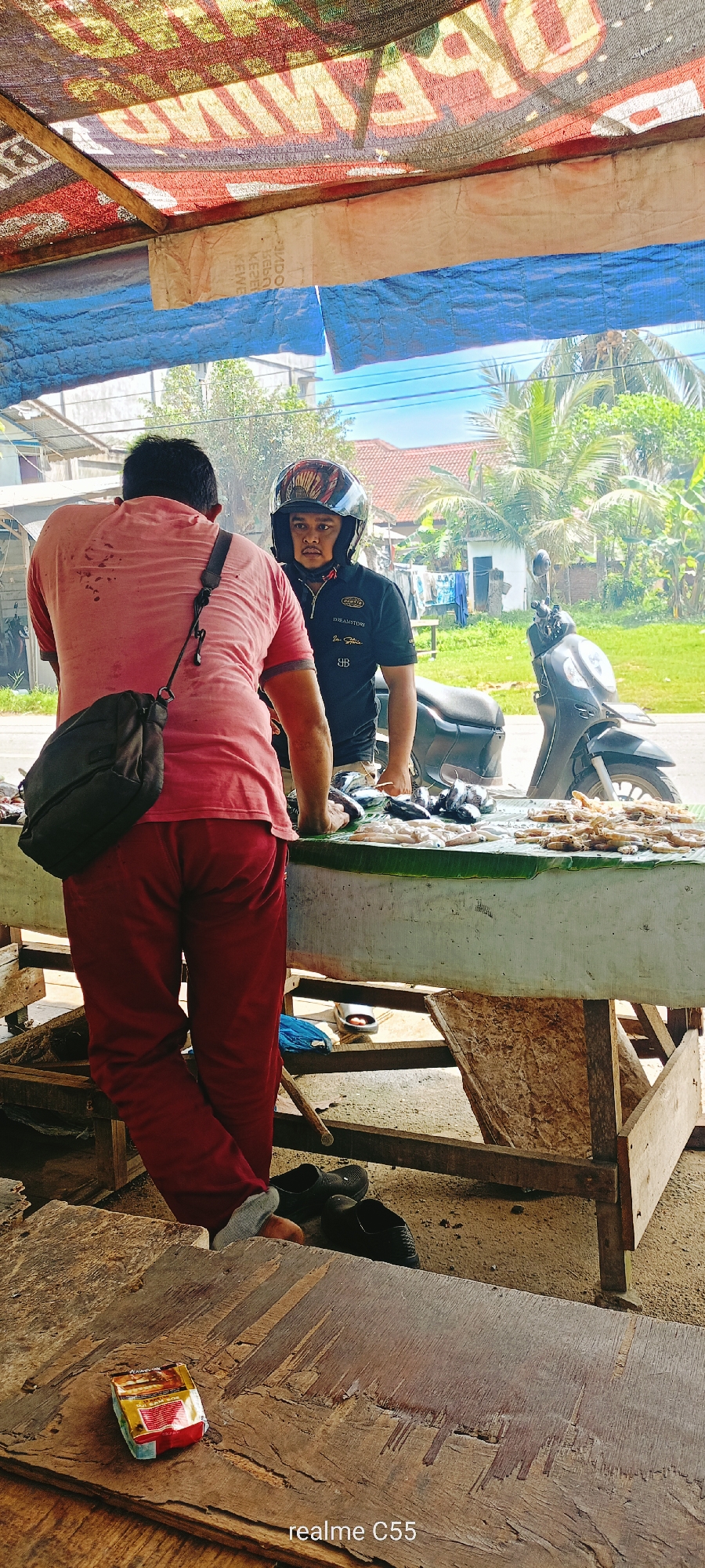 This image captures an outdoor scene in Lhokseumawe, Indonesia, depicting a roadside fish stall under an improvised canopy. In the foreground, a heavily textured wooden surface with visible grain and tool marks features a crumpled red and yellow package, possibly a cigarette pack, near the bottom left corner. A faint "realme C55" watermark is present at the bottom.
Centered in the midground, two adult males are engaged at a sales counter. The male on the left, facing away from the viewer, wears a faded red short-sleeved t-shirt with noticeable sweat stains on the upper back and loose-fitting maroon or dark red trousers. A black sling bag with a visible carabiner clip hangs over his right shoulder. He stands with his left foot slightly raised and resting against the wooden frame of the stall, wearing dark slip-on shoes.
The male on the right, facing towards the left, wears a black short-sleeved polo shirt with a zipper placket, a black full-face motorcycle helmet with a colorful patterned design, and a dark visor partially obscuring his face. Text on his shirt includes "DREAMSTORY," "I'm Shina," and "BB." He has a short beard or stubble visible around his chin. He stands behind a white, flat-surfaced display table, which appears to be a counter for selling goods. The surface of this counter is covered with green banana leaves, upon which various types of seafood are displayed for sale. These include silver-colored fish, smaller white fish, and piles of what appear to be shrimp or squid.
Behind the male in the helmet, a dark-colored scooter or motorcycle is parked. The background reveals an outdoor environment with lush green foliage, including palm trees, under a clear blue sky. Distant structures, including a building with a red-tiled roof and another with a white facade, are partially visible. Laundry appears to be hanging on a line near one of the distant buildings.
Above the scene, an overhead canopy provides shade. It consists of wooden beams supporting two layers of material. The upper layer is a patterned, possibly repurposed banner or tarp, displaying reversed text, notably "GEMILANG" and "SUPER" in red and yellow hues. Beneath this, a blue tarp and a piece of white fabric are visible, offering additional cover. The overall lighting suggests a bright, sunny day.