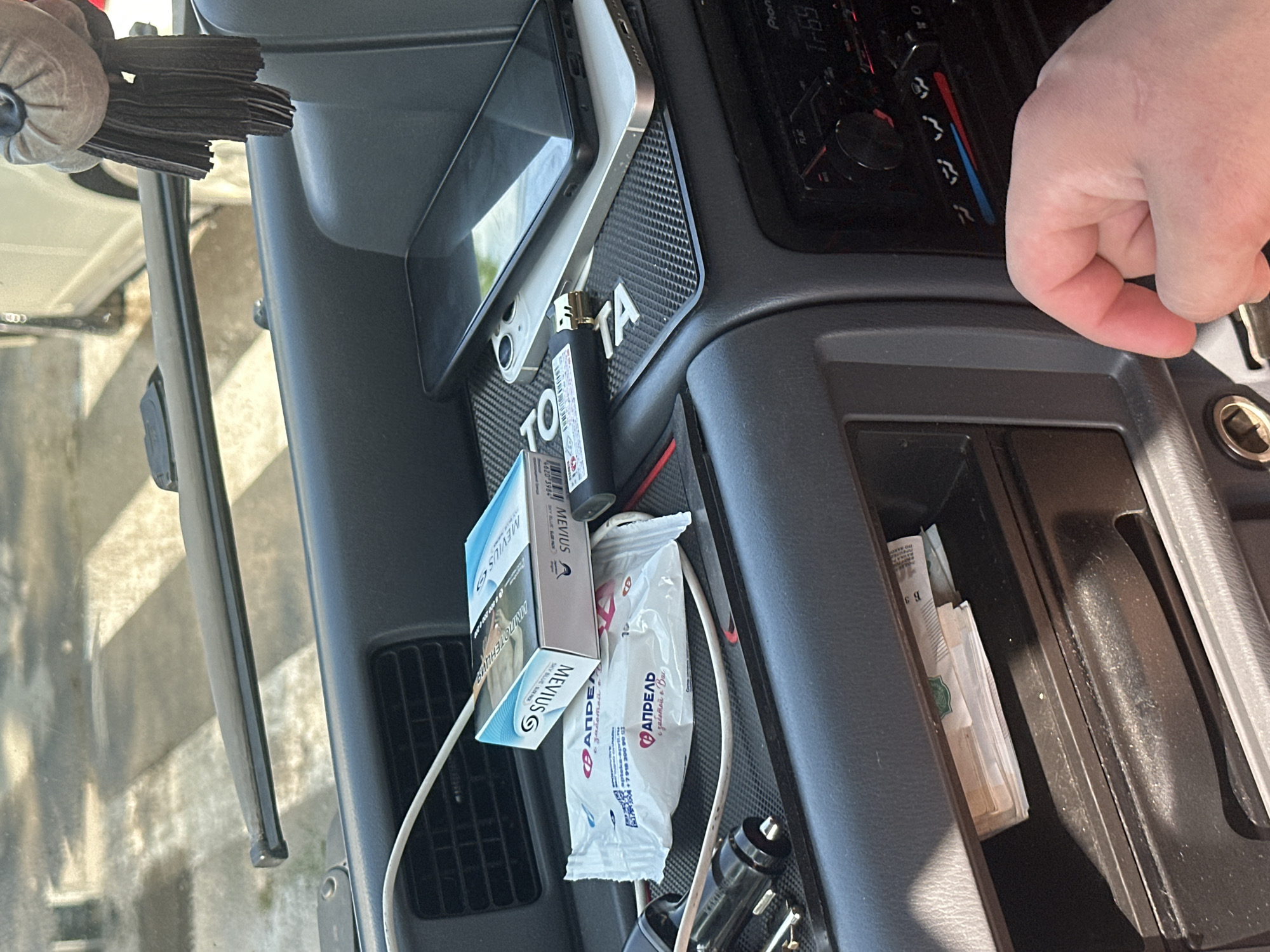 The image shows a close-up view of the interior of a car's center console. A hand, likely belonging to a person, is visible on the right side of the frame, reaching towards the console. Several objects are present: a pack of Mevius cigarettes, a lighter, two small white packets with text in Cyrillic that appears to be advertising some kind of product, a smartphone, and a stack of Russian rubles in the storage compartment. A car charger is plugged into a power outlet. The ambient lighting suggests it is daytime, but the interior of the car is dimly lit. The presence of Russian currency and text points to the location being Russia, specifically Birobidzhan as indicated in the prompt. The overall scene depicts common items found within a car's personal space. The text on the cigarette pack reads "MEVIUS SKY BLUE SONG" and other Russian words. The white packets have text including "АПРЕЛЬ" which translates to "April", and phrases like "заботой в Вас" meaning "with care for you". The car radio display shows "1-69".