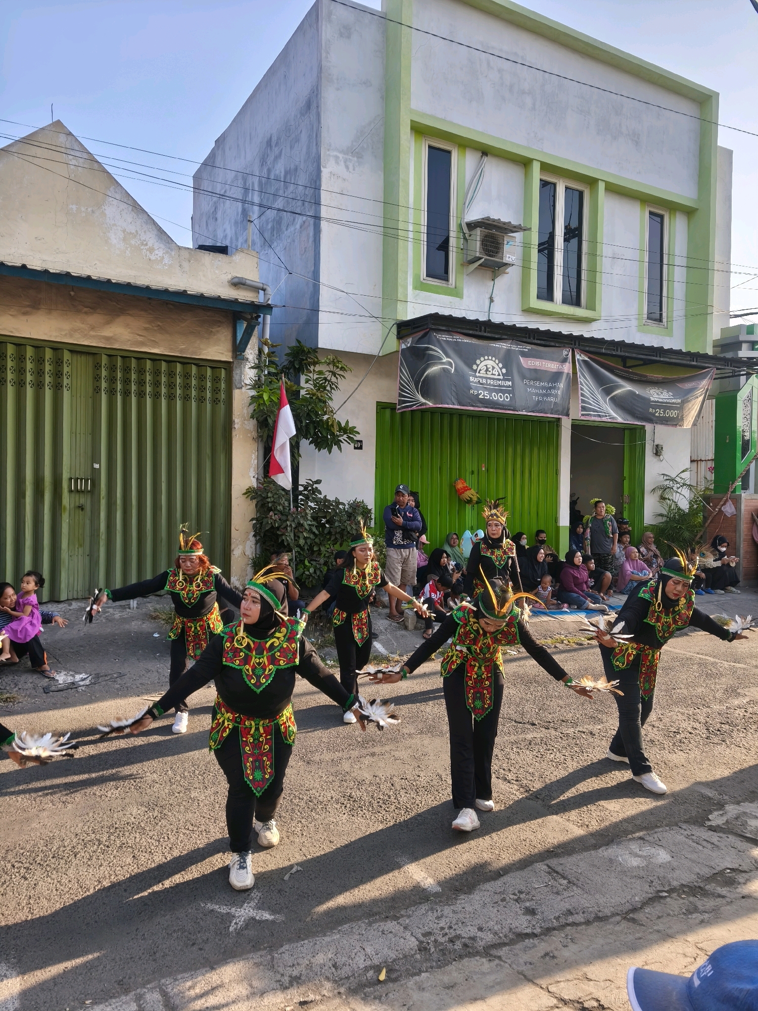 In Kepuh Kiriman, Indonesia, a group of six dancers in ornate, traditional costumes are performing a street parade. They are all wearing black pants and jackets adorned with colorful, embroidered patterns around the neckline and waist. Their headwear includes feathered headdresses. The dancers are holding feather props in each hand, and their movements suggest a choreographed performance. They are positioned on an asphalt road, casting long shadows from the bright sunlight.
Behind the dancers, a crowd of spectators is gathered on the side of the road, seated and standing. They are watching the performance, with a mix of adults and children. The background features buildings with green-trimmed windows and doors, and a large green folding gate. A few trees and foliage are visible, along with a flagpole flying the Indonesian flag. Banners with text and graphics are displayed above the storefronts. The overall scene captures a community event or festival taking place in a residential or commercial street.