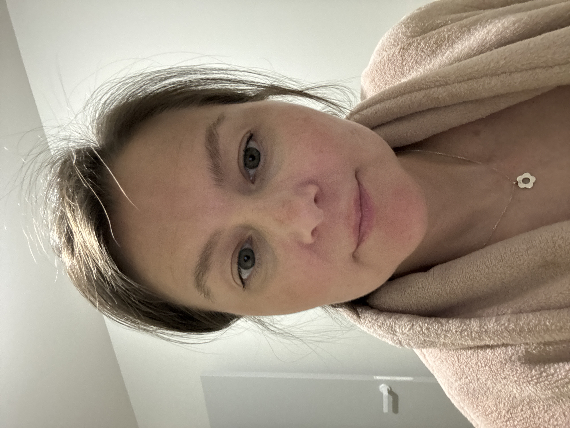 The image shows a close-up selfie of a person wearing a light-colored terrycloth robe. The person has fair skin, light brown hair, and green eyes. A delicate silver necklace with a flower-shaped pendant is visible on their chest. The background is a plain white wall. There is no indication of the city of Izhevsk, Russia, in the image.