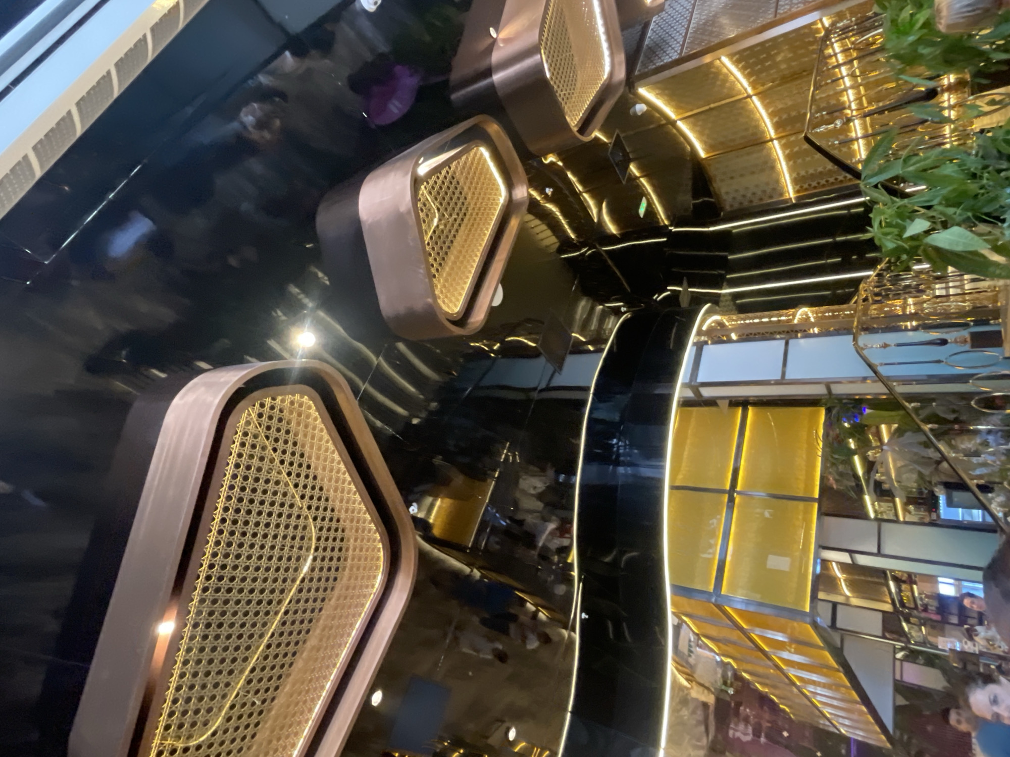 The image depicts an interior scene with a glossy black reflective surface dominating the foreground. Several decorative objects, possibly seating or planters, are arranged on this surface. These objects have a modern design, featuring rounded edges and a combination of copper-toned metal and woven cane-like material with a golden hue. Integrated lighting illuminates the cane sections, creating a warm glow.
In the background, more of the reflective black surface is visible, along with architectural elements that suggest a stylish, contemporary indoor space. There are reflections of lights, what appear to be plants, and hints of people. The overall aesthetic is luxurious and high-tech, with a focus on reflective materials and strategic lighting. The scene appears to be well-lit, suggesting it's daytime or the interior lighting is designed to be very bright. No specific activity is clearly discernible, but the presence of people in the reflections indicates it is an occupied space. The context of Barcelona, Spain, does not provide any overt visual cues within the image itself, but suggests an urban, possibly commercial or entertainment venue.