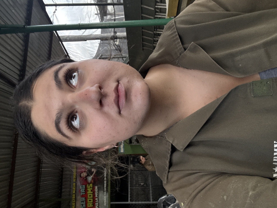 A woman is captured in a close-up, slightly angled self-portrait. Her dark hair is pulled back, highlighting her face with prominent eyebrows and lashes. She is wearing a green, collared shirt that appears to be a military-style jacket. In the background, there is corrugated metal roofing and hints of an outdoor setting. The overall image has a casual, selfie-style aesthetic.