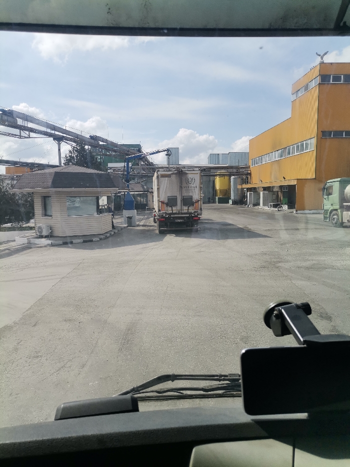 The image, captured from the interior of a vehicle in Karapelit, Bulgaria, depicts an industrial complex yard under a partly cloudy sky. The foreground reveals a vehicle dashboard with windshield wipers and a black mounting bracket on the right.
In the mid-ground, a white commercial truck with a box trailer is positioned centrally, facing away from the viewer. The rear of its trailer bears the inscription "PREMIER PLACE" and is fitted with a Bulgarian license plate (e.g., E 2110 MA). Faint dust or exhaust trails are visible behind the truck, suggesting recent movement. To the left of the truck, an octagonal security booth or guardhouse with a brown, multi-faceted roof and an external air conditioning unit is situated next to a paved area with a low curb.
Above and behind the security booth, an extensive network of metal structures, including conveyor belts or piping, spans across the upper-left and central portions of the frame, indicating active industrial processing. Green foliage is visible beneath these structures. To the right, a prominent multi-story industrial building with yellow-orange cladding and horizontal rows of windows stands. A bird-shaped sculpture or weather vane is affixed to the building's rooftop corner. Further to the right, the front section of a green tanker truck is partially visible. The ground throughout the yard is paved with concrete or asphalt and appears somewhat dusty. No individuals are visible within the scene.