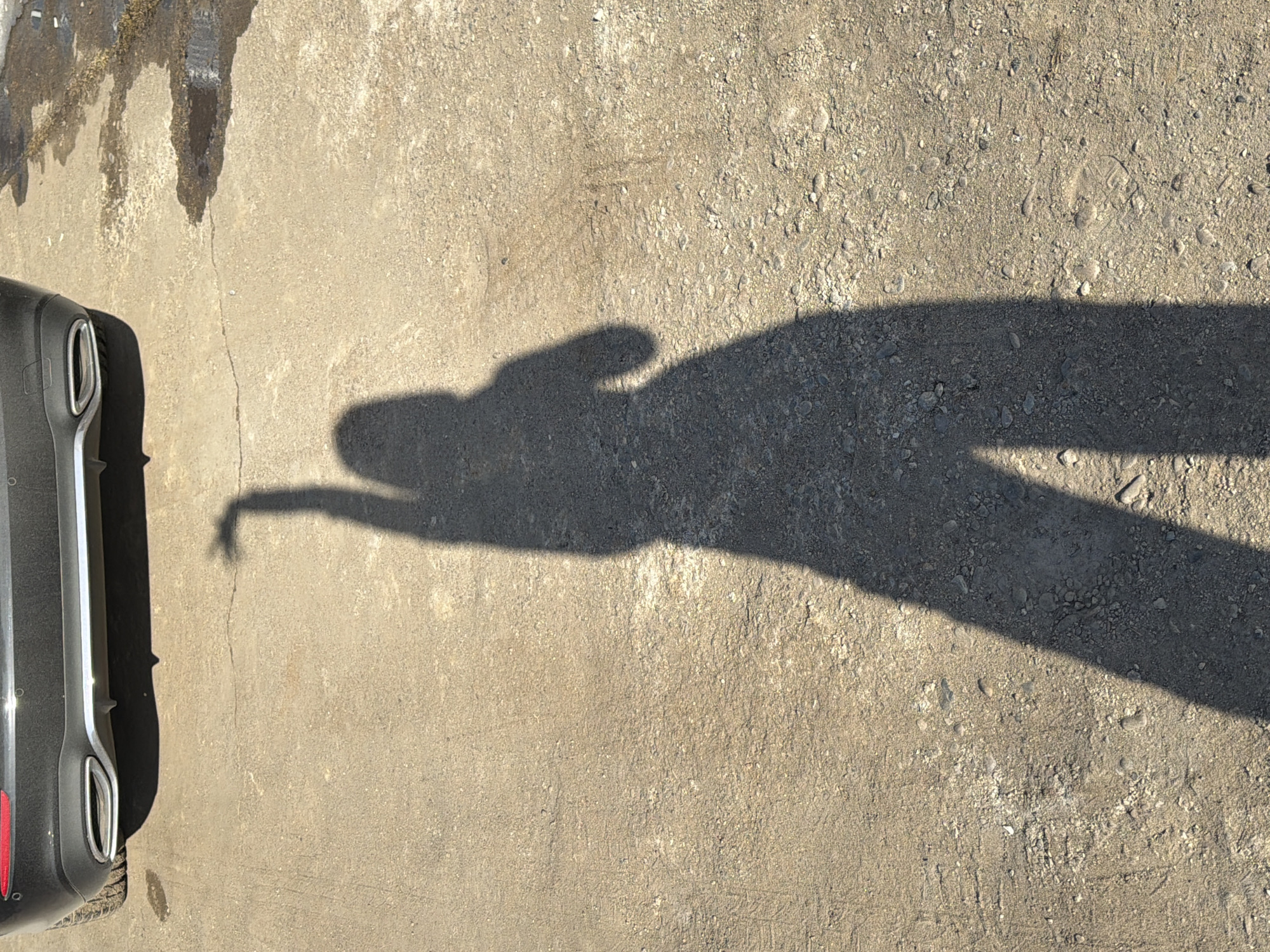 The image shows a section of pavement or concrete, likely outdoors. A portion of a dark-colored vehicle is visible on the left side of the frame. A shadow of a person is cast across the pavement. The pavement appears to be uneven, with some areas appearing smoother than others. Based on the details and context of the image, it appears to be taken outside, likely in a parking lot or near a roadway. There is no verifiable information to place this scene in Chaikovsky, Russia, or other cities.