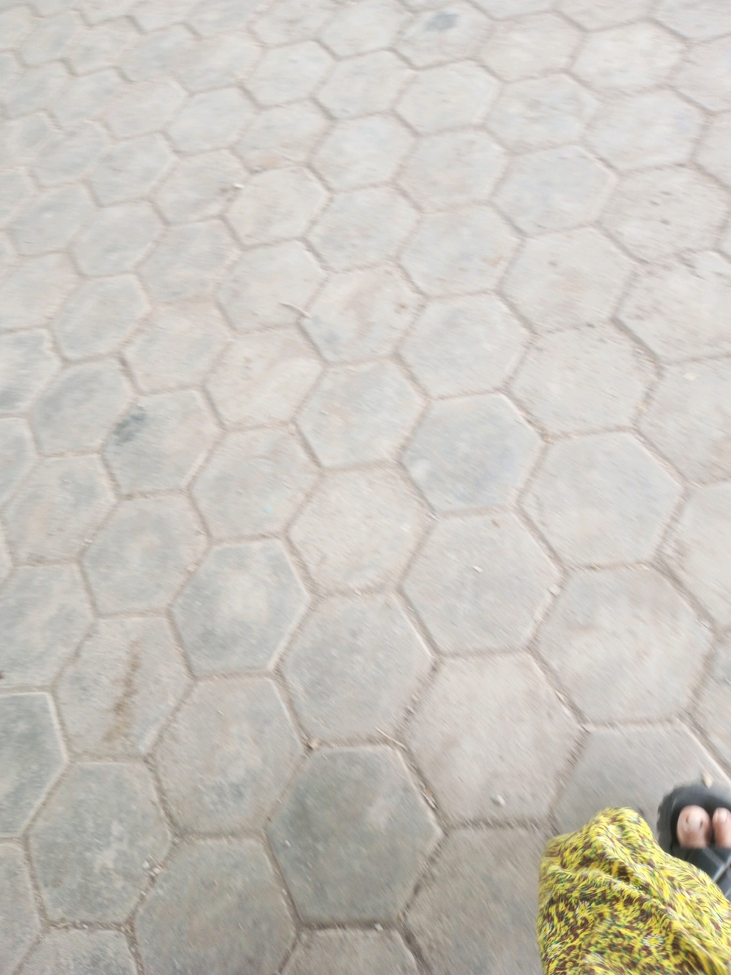 A person wearing a yellow and green patterned garment and black sandals is visible in the lower right corner of the image. They are standing on a ground covered with hexagonal concrete pavers. The pavers are light gray and are arranged in a repeating pattern. The overall scene appears to be an outdoor setting in the city of Kalibeber, Indonesia, possibly a street or walkway.