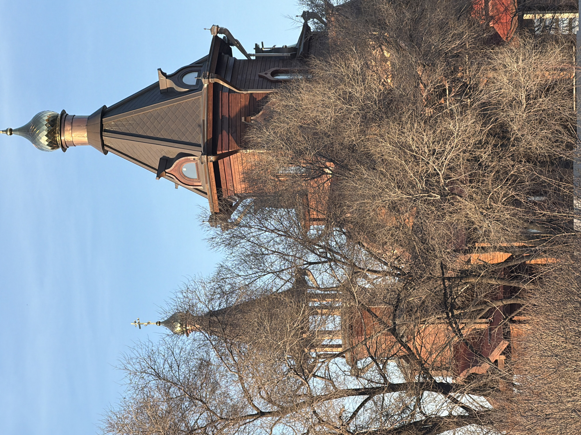 A low-angle shot shows the top of St. Nicholas Church in Krasnoyarsk, Russia. The church has two onion-shaped domes with crosses on top. The roofs are reddish-brown and black. Bare trees surround the building. The sky is light blue.