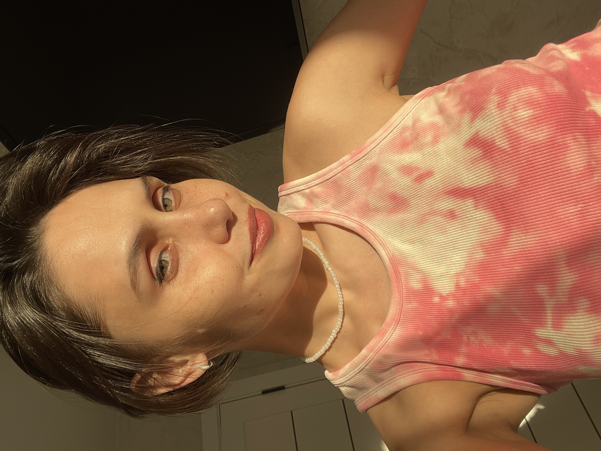 A woman is pictured in a well-lit interior space. She is wearing a pink and white tie-dye ribbed tank top and a white beaded necklace. The background includes a dark surface, possibly a wall or ceiling, and a white door. The location in Izhevsk, Russia, is not evident from the image.