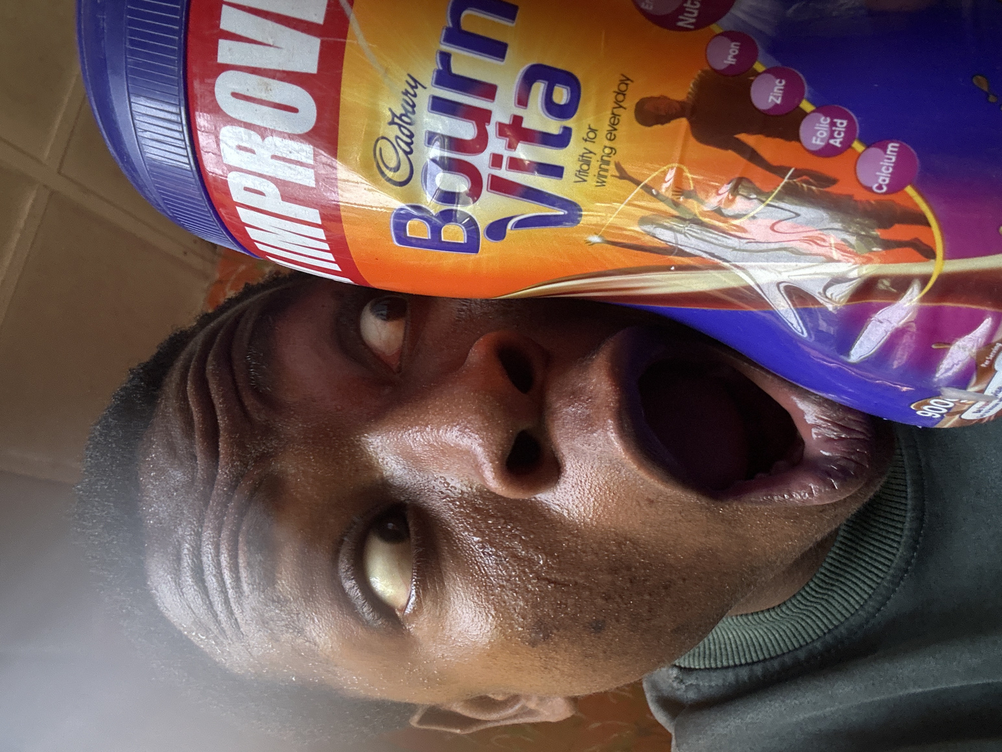A close-up, rotated portrait shows a person with dark skin holding a large Cadbury Bourn Vita container directly over their face. The person has an exaggerated expression, with wide eyes and a mouth gaping open as if about to consume the product directly from the tub. Their forehead shows prominent wrinkles, and their skin appears shiny, possibly with sweat or oil. They are wearing a dark green or gray crew-neck top.
The purple and orange Bourn Vita container features the brand name "Cadbury Bourn Vita" and the slogan "Vitality for winning everyday." Visible nutritional elements listed on the packaging include "Iron," "Zinc," "Folic Acid," and "Calcium," along with partially visible words like "IMPROV" (likely "IMPROVED"), "Nut" (for nutrition), and "Er" (for energy). A "900" (likely grams) marking is also visible, along with a partial "Per Serving." The background is indistinct, suggesting an indoor setting. The image conveys a sense of humor or exaggerated eagerness.