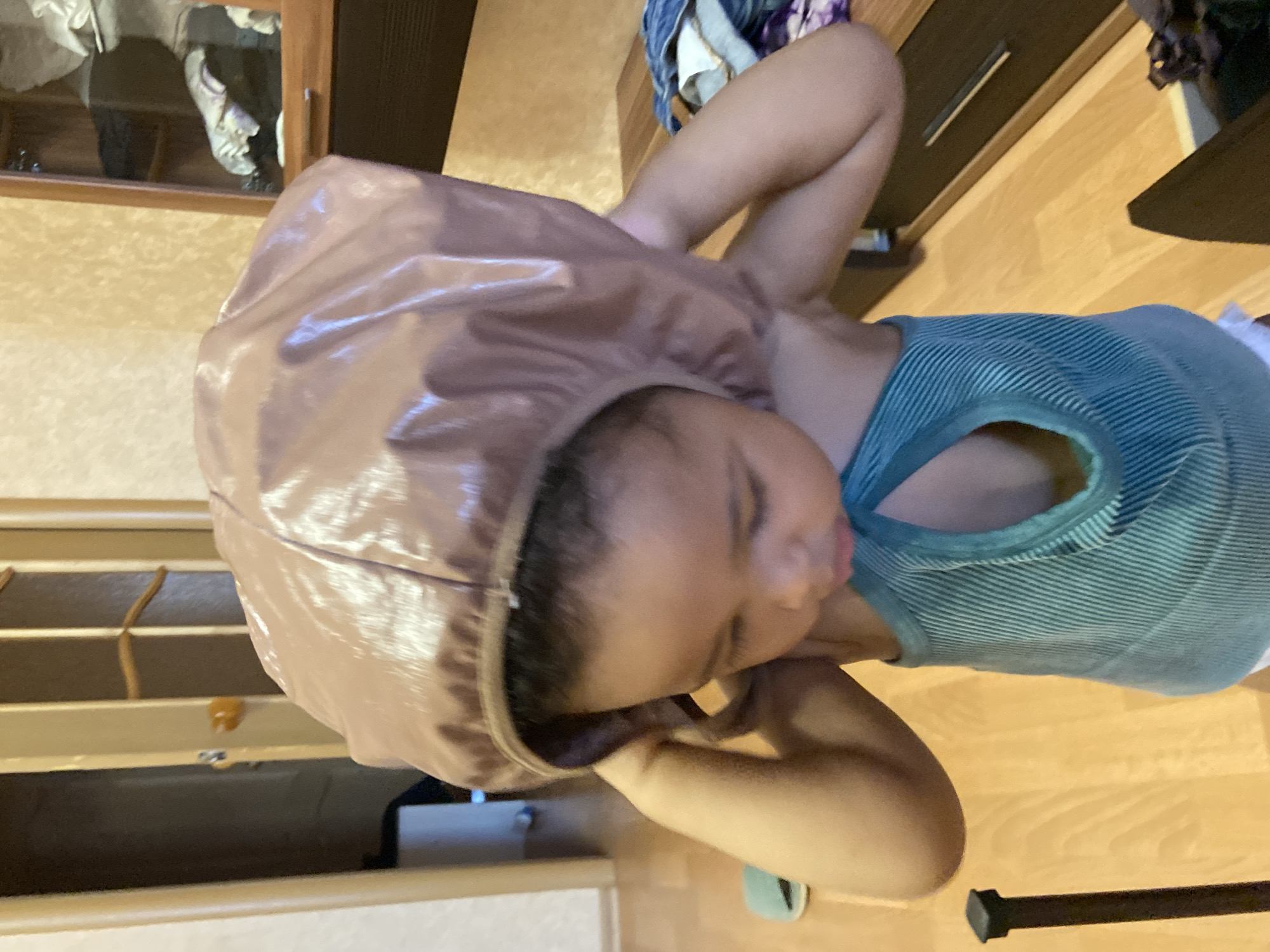 A young individual with a brown bonnet is lying down indoors. The person wears a blue and white striped tank top. The environment includes wood flooring, furniture, and what appears to be storage cabinets. The specific location within Kazan, Russia, cannot be determined from the image.