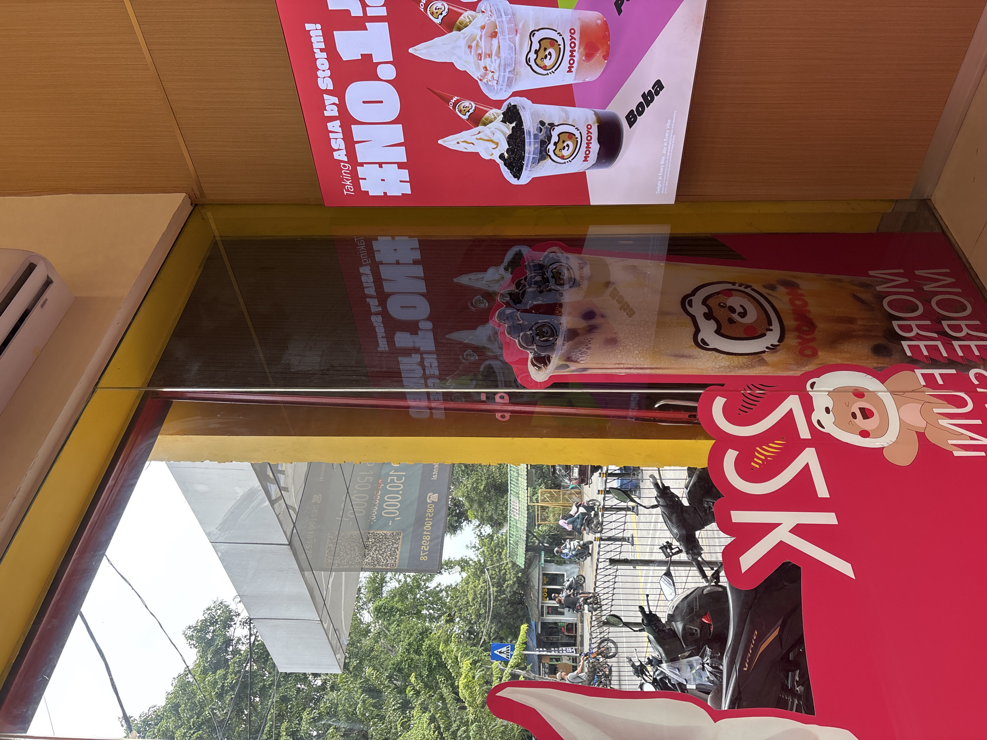 The image depicts the storefront of a MOMOYO bubble tea shop located in North Jakarta, Indonesia. The shop has a glass front with promotional stickers and posters. One poster features MOMOYO bubble tea and an ice cream product. A second advertisement highlights a "55K" special offer with a cartoon bear character on a pink background. Reflections in the glass show the street outside, including trees, parked motorcycles, and a crosswalk. The surrounding buildings are visible in the reflection, including businesses with signage.