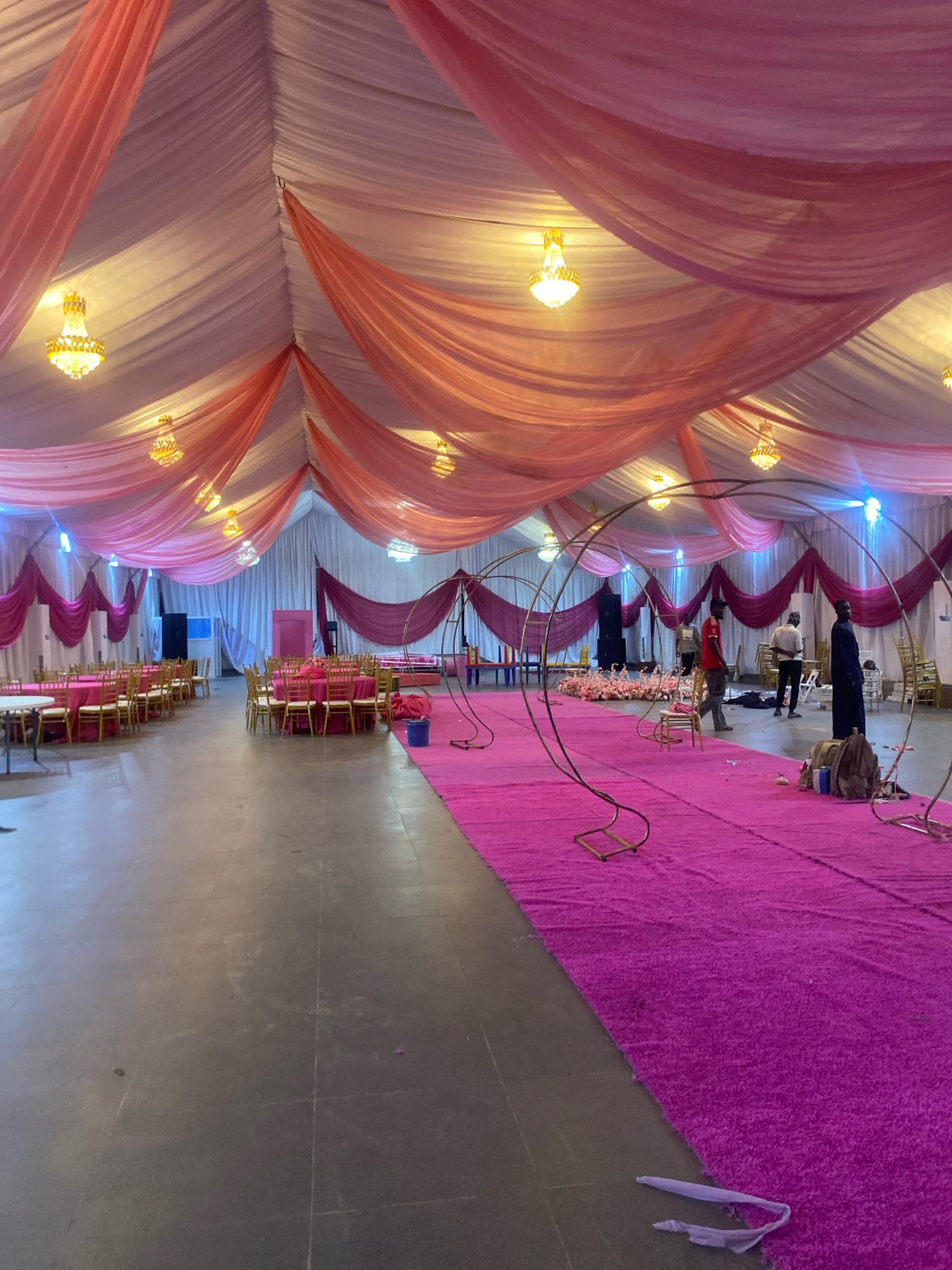 An indoor event hall is set up for a celebration, likely a wedding or party, within Barden Yana, Nigeria. The hall is draped with white and pink fabric, creating a canopy effect with chandeliers hanging from the ceiling. A bright pink carpet runs down the center of the room, leading towards a large floral arrangement and decorative circular arches. Seating arrangements consist of round tables covered in pink tablecloths and surrounded by gold chairs. Several individuals are present, some appearing to be setting up the event, while others are standing and observing the preparations. The overall atmosphere suggests an ongoing event setup.