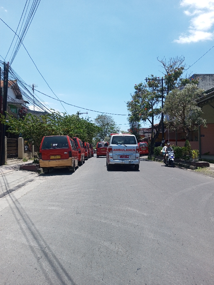 The scene is depicted on an asphalt street in the city of Tagtse Tashidingkha, Bhutan, under a clear blue sky with sparse clouds. Overhead utility lines crisscross the upper portion of the frame.
On the left, a series of red vans are parked curb-side. The nearest red van has a tan lower rear panel, a partially visible yellow license plate displaying "F 7979 YB", and a "07" sticker on its rear window. Behind the parked vehicles are a sidewalk, a dark fence, dense green foliage, and residential structures.
In the middle of the street, a white ambulance van, marked with "AMBULANCE" in blue text and a red and white checkered stripe, faces away from the viewer. Its license plate reads "F 1421 MZ".
To the right of the ambulance, a single individual rides a white scooter, also moving away from the viewer. The rider wears a light-colored shirt. The right side of the street features a sidewalk, a utility pole, various trees with green foliage and some reddish blooms, and more buildings in the background. The asphalt road shows minor surface wear.