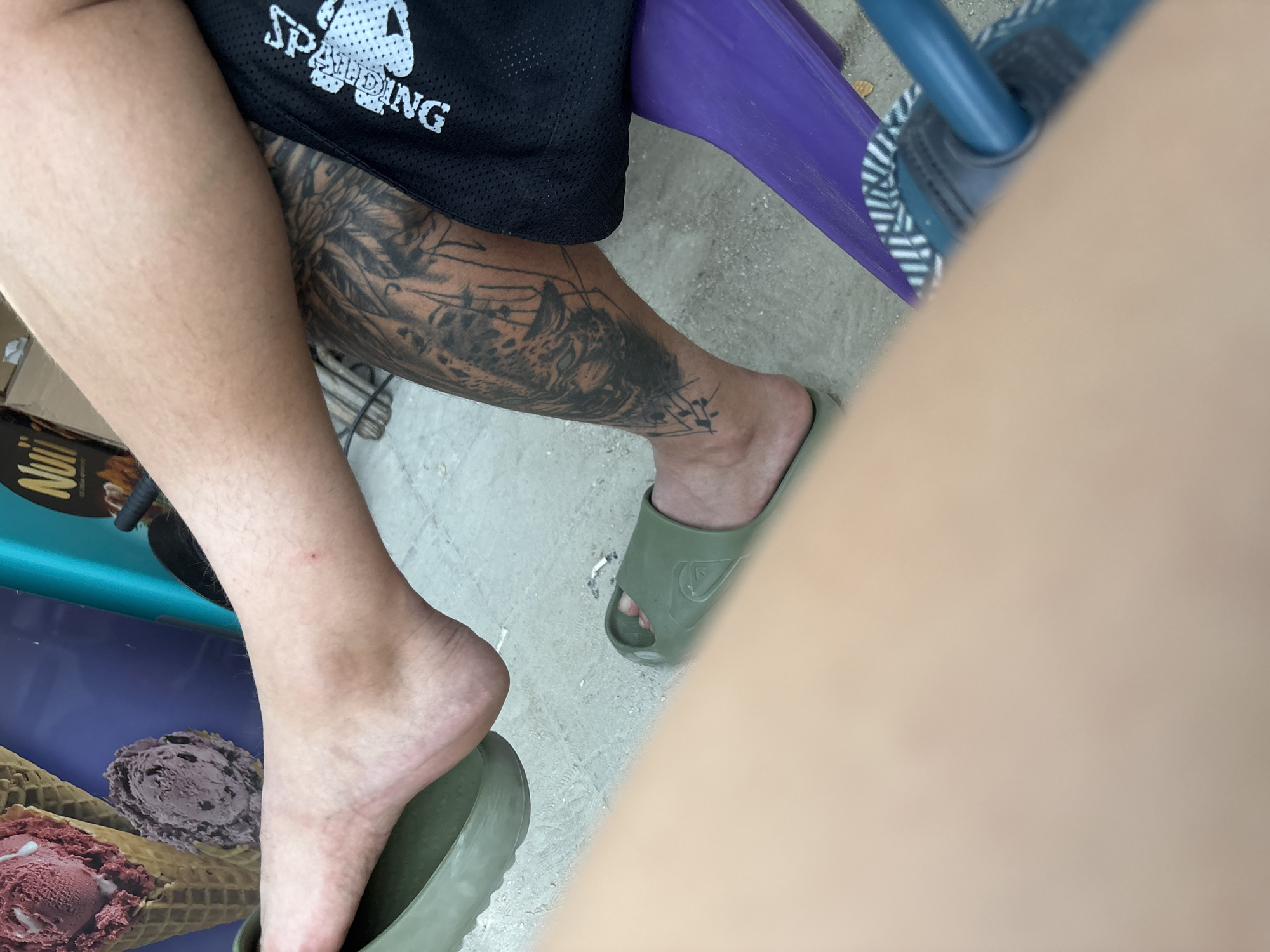 This outdoor scene, located in Kostievo, Bulgaria, depicts the lower extremities of a person, likely in a seated position. The person has fair skin with visible body hair on the upper calf. The left foot is bare and partially rests on an olive green slide with a textured sole. The right foot is fully inserted into a matching olive green slide, which features an embossed logo on the strap. A prominent black-ink tattoo, depicting an intricate design of what appears to be an animal head (possibly a tiger or similar feline) surrounded by geometric lines, covers the lower right leg from below the knee to above the ankle. Black athletic shorts with a white "Spalding" logo are visible on the right thigh. The ground consists of light-colored concrete or compacted sand, on which two discarded cigarette butts are present. In the background, a commercial poster displays two ice cream cones, one with a scoop of red ice cream and another with a scoop of purple or dark brown ice cream, alongside the brand name "Nuii." A segment of a purple object, possibly a chair leg or a small stool, is visible in the lower right, partially covered by a blurry light brown object in the immediate foreground, which is likely another person's limb.