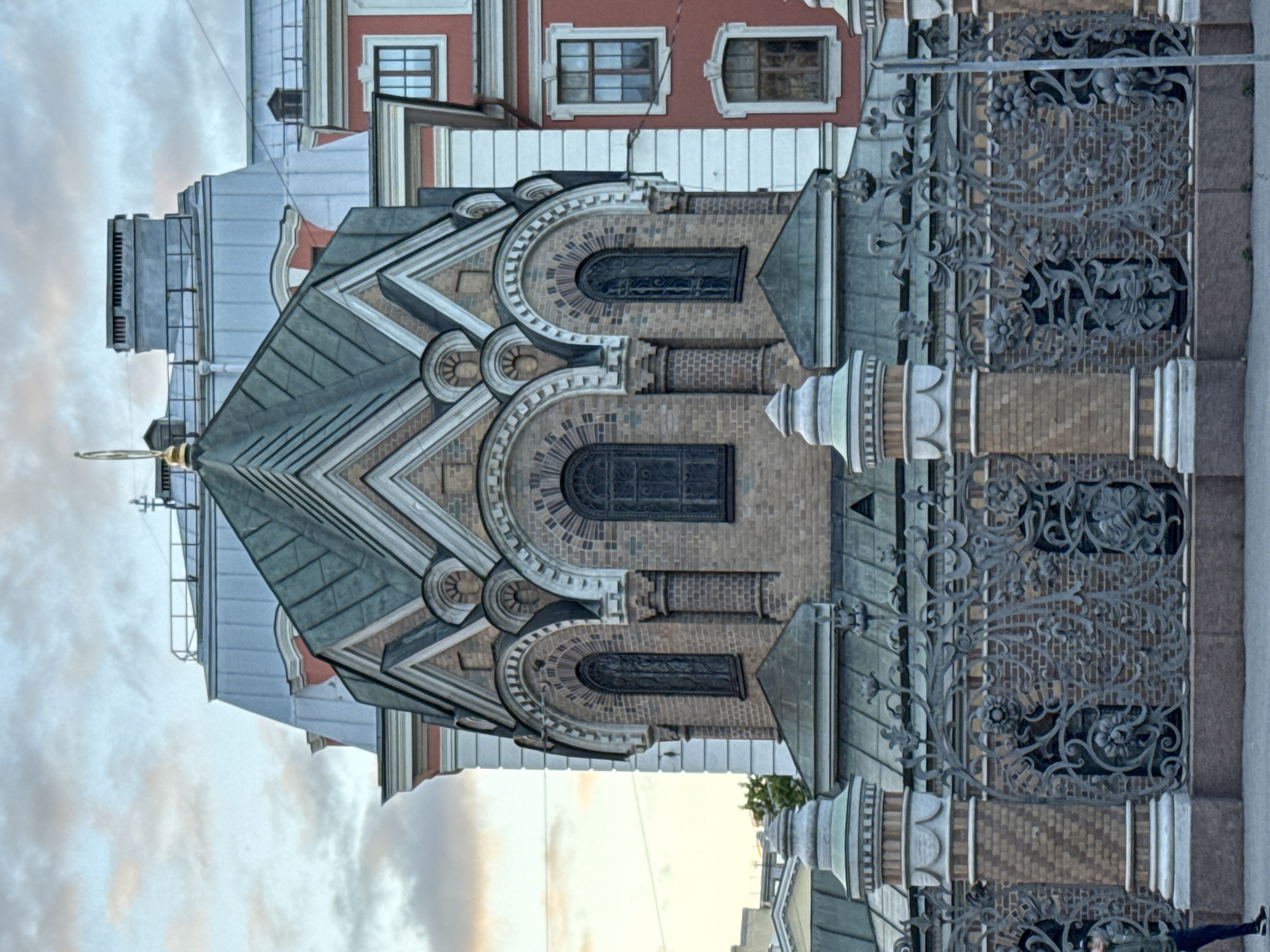 This image shows the Church of the Resurrection of Christ, also known as the Church of the Savior on Spilled Blood, located in Saint Petersburg, Russia. The exterior of the church is visible, featuring intricate brickwork, arched windows, and a green domed roof. In the foreground, an ornate wrought-iron fence with floral motifs separates the church from a sidewalk. A person in a green jacket and dark pants is partially visible on the left side of the frame, walking on the sidewalk. The sky above is cloudy with hues of pink and orange, suggesting either sunrise or sunset. The church's detailed architecture and decorative elements are prominent in the composition.