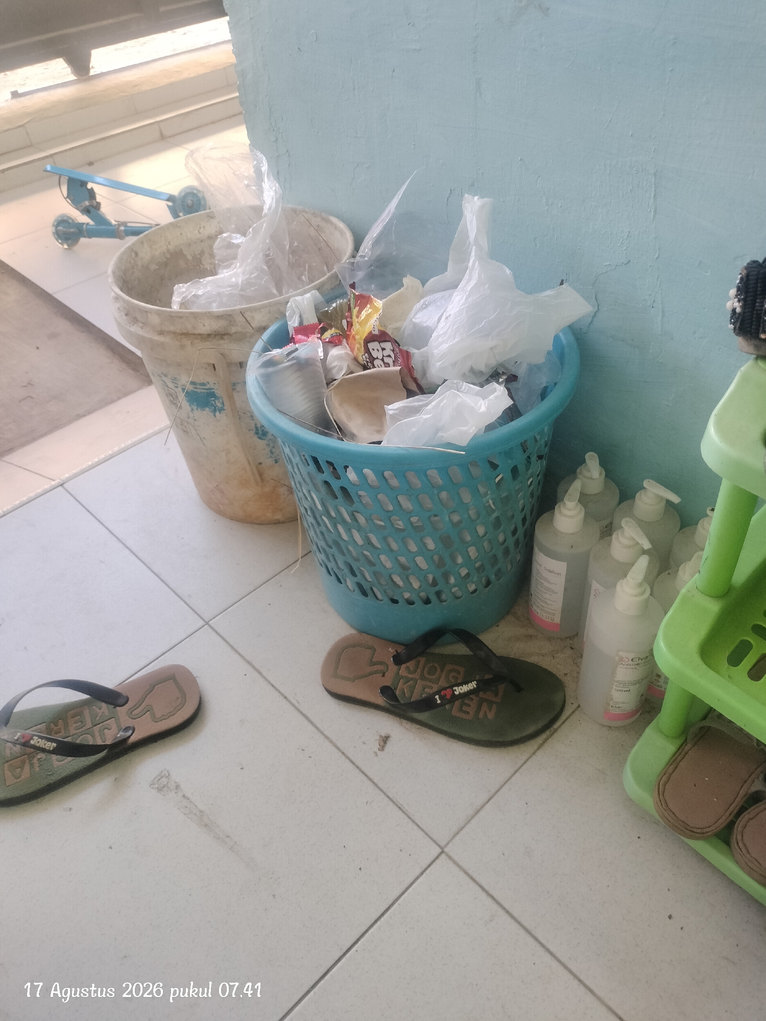 A cluttered outdoor scene, likely a side area or walkway, is captured in the image, dated August 17, 2026, at 07:41. The ground is tiled in a light color with visible grout lines. On the left, a dirty, off-white bucket contains plastic bags and other refuse. Next to it, a blue plastic basket is overflowing with similar waste. To the right of the basket, several clear plastic bottles with white pump dispensers are arranged, some appearing to be hand sanitizer or cleaning solutions. One bottle is labeled "Elva Antiseptik". A multi-tiered green plastic shelf unit stands to the far right, holding a pair of brown flip-flops on the bottom shelf. Two pairs of flip-flops are on the tiled floor; one is dark with a "Joker" logo, and another, partially visible, is a lighter brown. In the background, a blue child's scooter with three wheels is propped up. The wall behind the objects is a light blue, appearing to be an exterior wall. The presence of the scooter and hand sanitizer suggests this might be a residential or a communal outdoor space, possibly in a hotter climate given the open walkway and scooter. Given the visible timestamp and the common usage of such items, it is plausible this scene is located in the city of Kupang, Indonesia, a city known for its warm climate and residential areas.