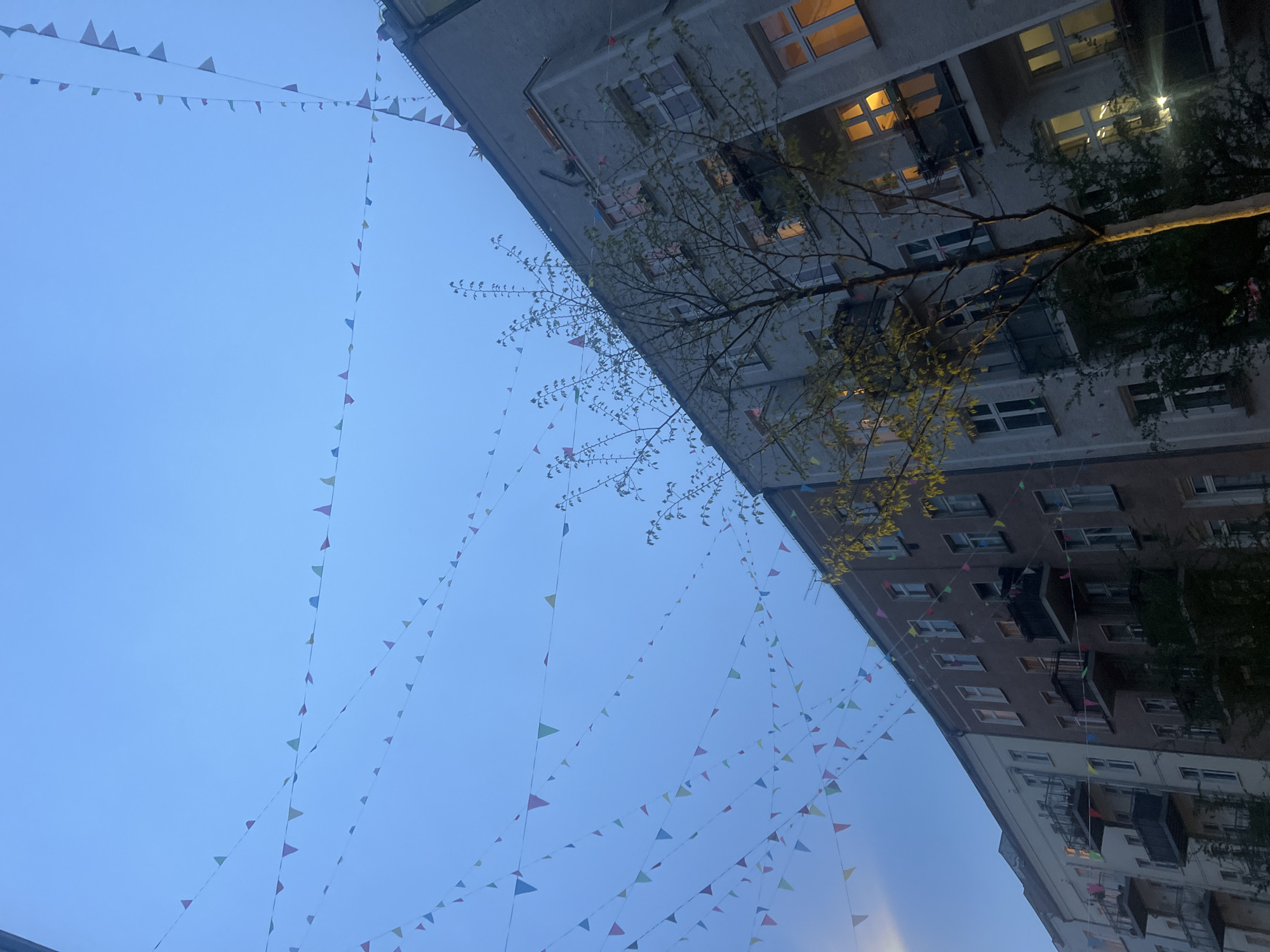The image shows a view looking up between two buildings in Berlin, Germany. Colored triangular flags are strung between the buildings, creating a festive atmosphere. A tree with budding leaves stands in front of the buildings, partially obscuring their facades. The buildings appear to be residential apartments with windows, some illuminated. The sky is a light blue, indicating either early morning or late evening.