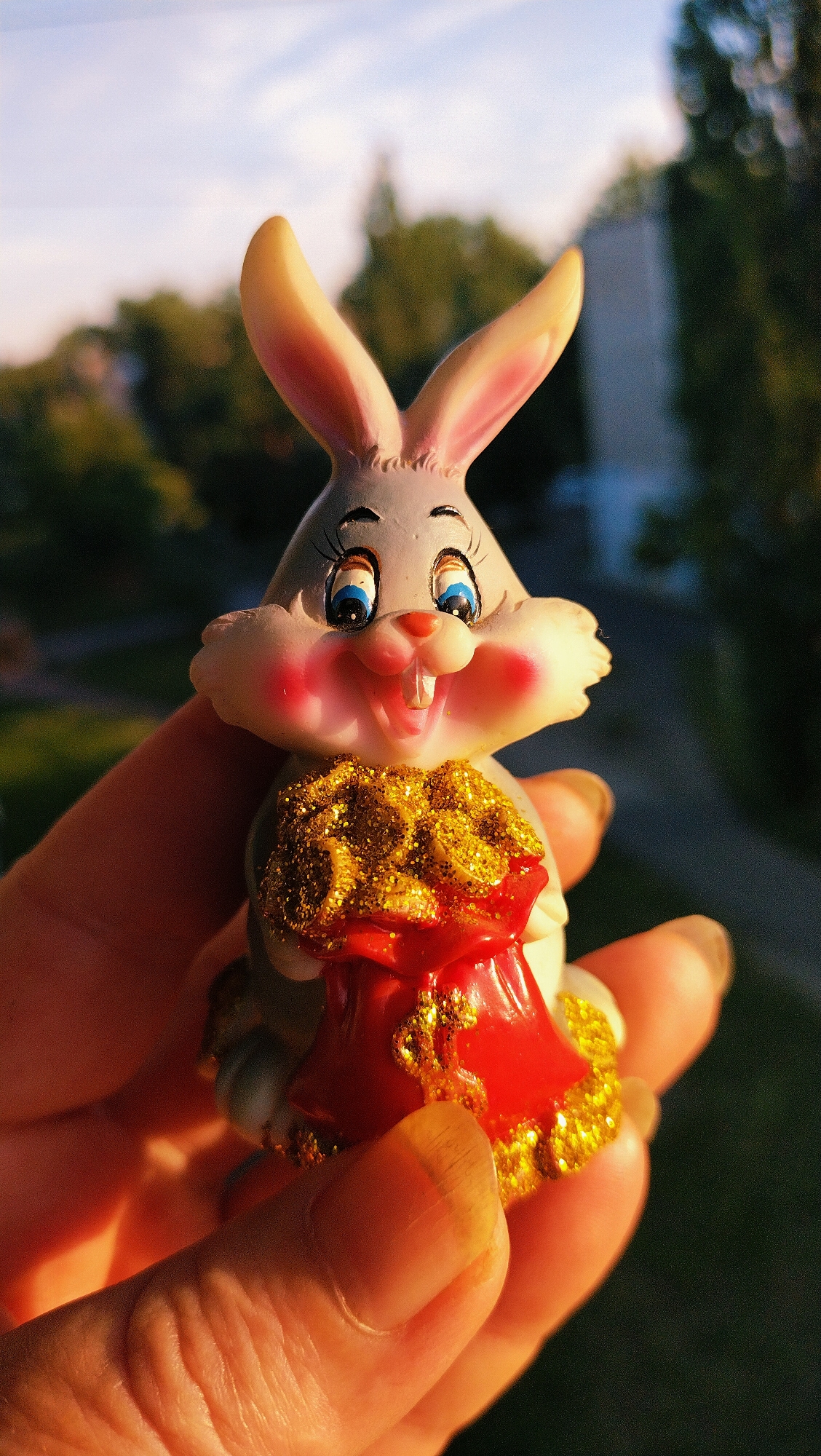 A close-up shot shows a person's hand holding a small, grey bunny figurine with large blue eyes and rosy cheeks. The figurine is wearing a red and gold sparkly outfit, possibly a dress or a sack. The background is blurred, showing trees and a building, suggesting an outdoor urban setting, possibly in the city of Horishni Plavni, Ukraine, given the context. The lighting indicates it's either sunrise or sunset, with the sun casting a warm glow on the figurine and hand.