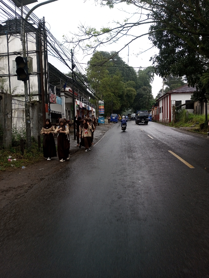 The image presents an overcast, wet street scene. On the left side of the asphalt road, a group of approximately seven to nine young females, appearing to be students, walk along a dirt shoulder. They are dressed in uniforms consisting of dark brown long skirts or trousers, light-colored long-sleeved tops, and several wear brown headscarves (hijabs). They carry bags. The road surface is visibly wet, reflecting the gray sky. A faded yellow dashed line is present on the road.
A motorcycle with a rider is positioned in the middle of the road, traveling away from the viewer. Further in the distance, a dark-colored truck and multiple blue microvans are visible.
To the left of the road, a concrete fence separates the street from a multi-story white building. A utility pole heavily laden with a dense network of black electrical and communication cables supports a black, vertically oriented traffic light with three lamps. Beyond this, a series of commercial buildings or shopfronts are visible, with clothing and other items displayed. A colorful advertising banner is also present.
On the right side of the road, a single-story white building with prominent vertical red stripes and a reddish-brown tiled roof stands. Lush green trees and dense foliage border the scene, particularly on the right and in the background. The environment is urban or semi-urban under cloudy conditions.
The visual characteristics, including the specific style of school uniforms, architecture, and vehicles, are inconsistent with the specified location of YonPhoogla, Bhutan, and are more characteristic of a Southeast Asian country like Indonesia or Malaysia.