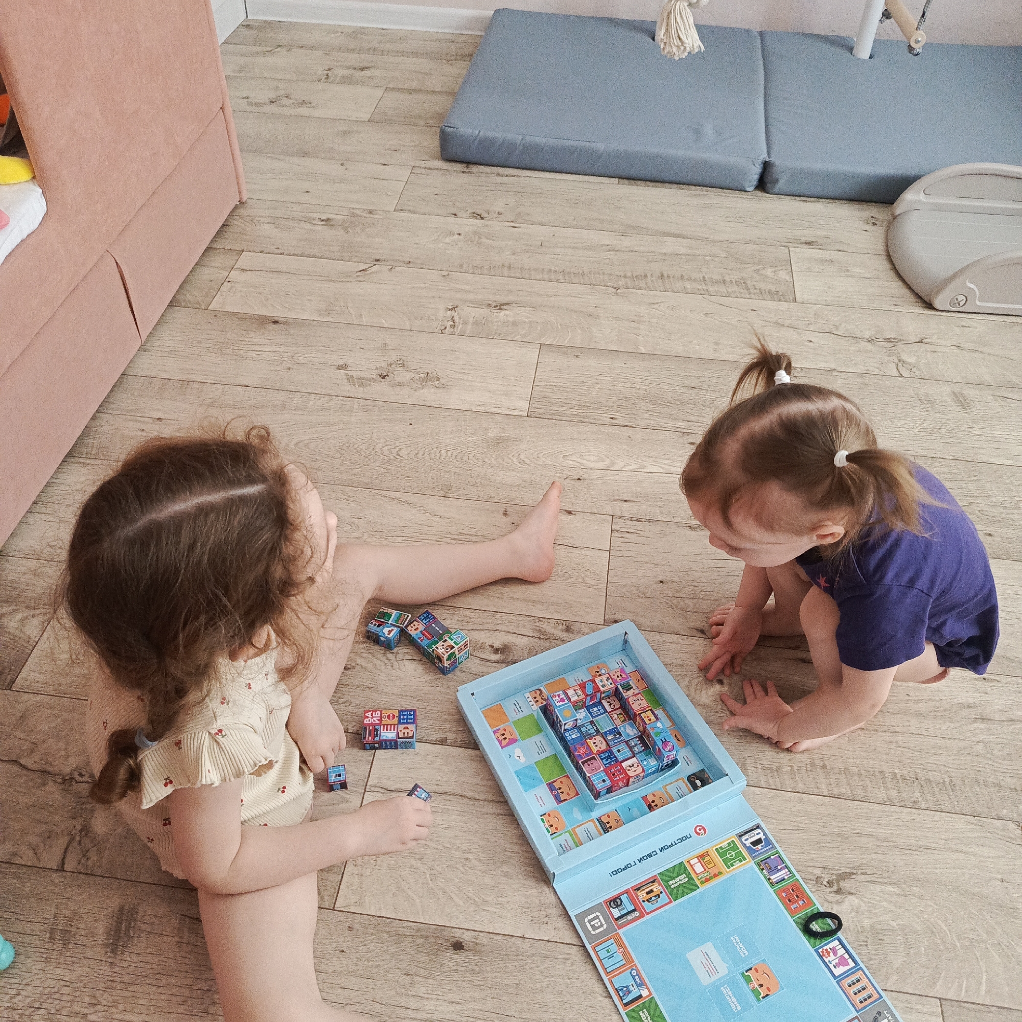 Two young girls are sitting on a wooden floor, engaged in playing a building block game. One girl, with dark hair styled in a braid, is wearing a light-colored patterned romper and is partially extended, with one leg stretched out. The second girl, with dark hair in pigtails, is wearing a dark blue t-shirt and is crouched down. They are both focused on a blue rectangular box, which contains an assortment of building blocks, some of which have been assembled into structures. Additional blocks are scattered on the floor near the girls. A section of a pink sofa is visible in the background, along with a blue mat. There is light wall color with no distinctive wall hangings. The image was taken in an unidentified room in the city of Марковский, Russia.
