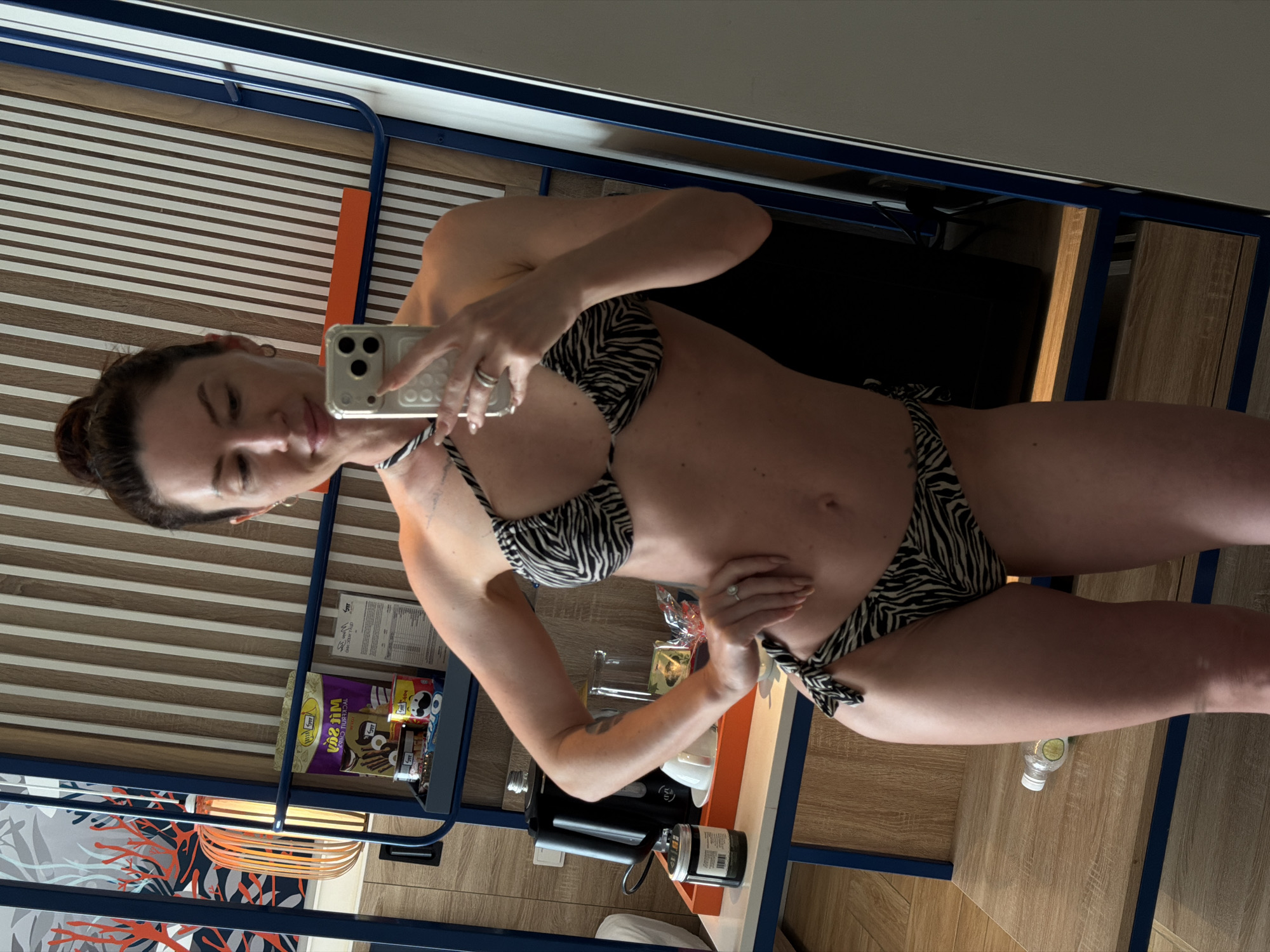 A woman in a zebra-print bikini stands in a room, taking a mirror selfie with her phone. The room appears to be a hotel room, with a bed, a shelf with toiletries and snacks, and a wooden wall. The woman has her hair in a bun and is wearing rings on her fingers. The image is taken from the waist up, showing her torso and arms.