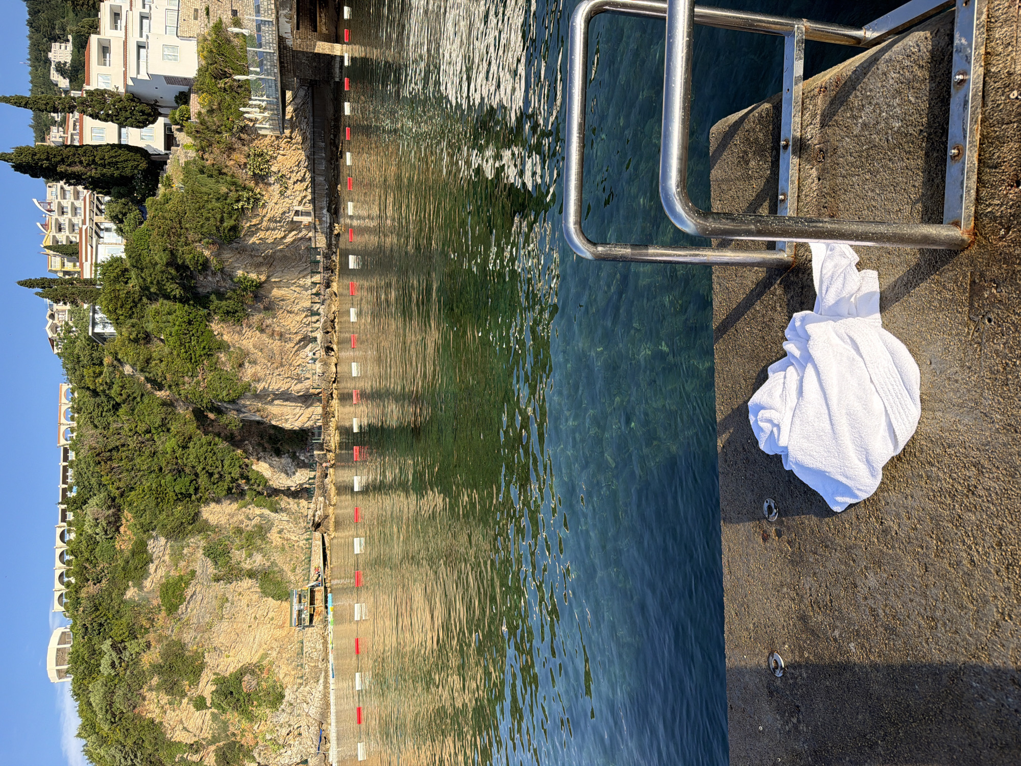 This image shows a beach scene in Budva, Montenegro. A rocky cliff with trees and buildings rises along the coast. A concrete pier juts into the water, with a stainless steel ladder extending down to the water. A white towel rests on the pier next to the ladder. The water is clear, with reflections of the surrounding landscape on the surface. Red and white floating buoys border the swimming area.