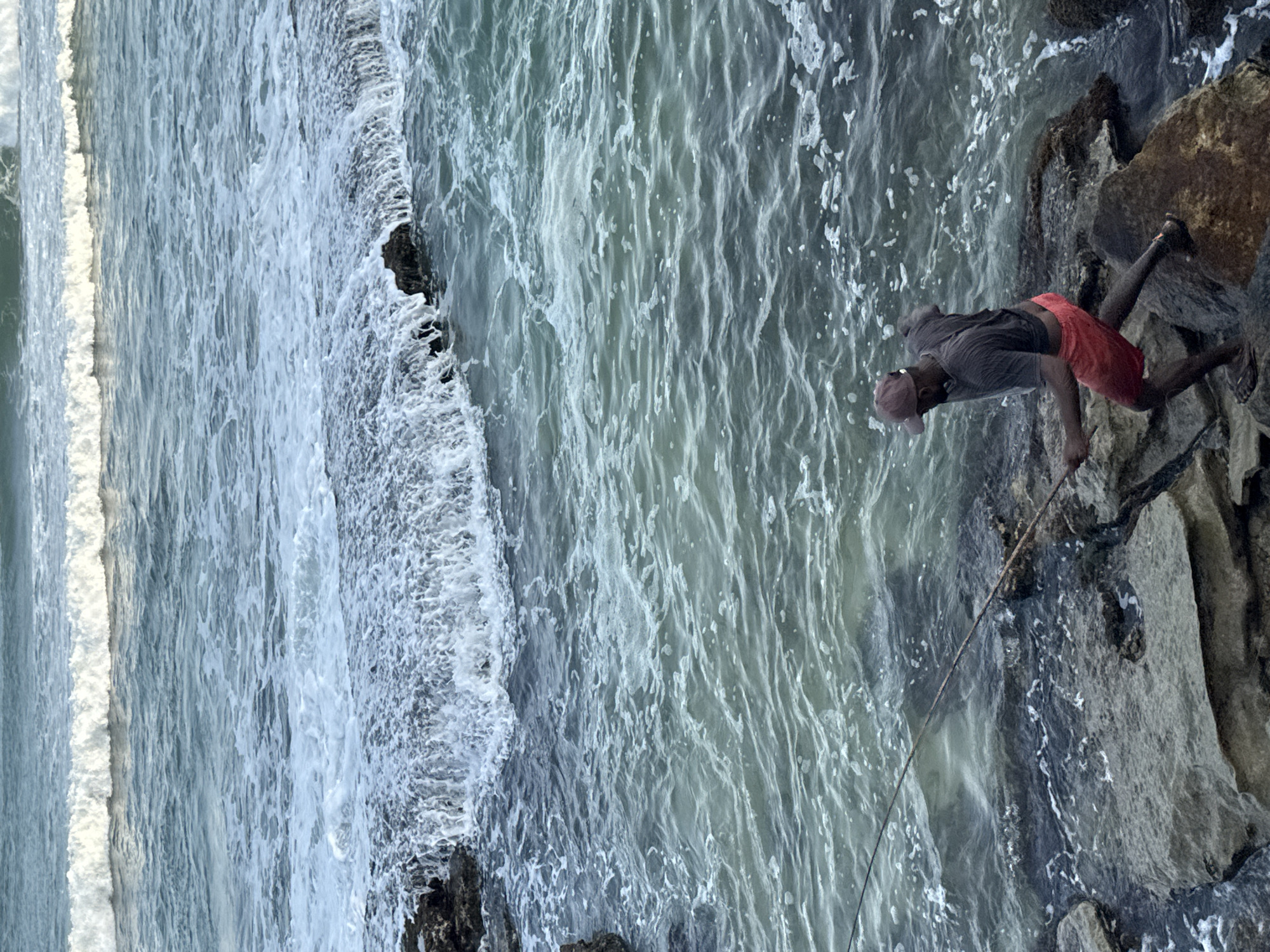 A man fishes from the rocky shore of Goviyapana, Sri Lanka, with waves crashing against the rocks. The man is wearing a maroon baseball cap, a grey t-shirt, and red shorts. He is holding a fishing rod and appears to be in mid-action. The water is a deep blue-green, with white foam from the waves. The rocks are dark and jagged, providing a natural breakwater. The scene is captured from a slightly elevated perspective, looking down at the man and the ocean.