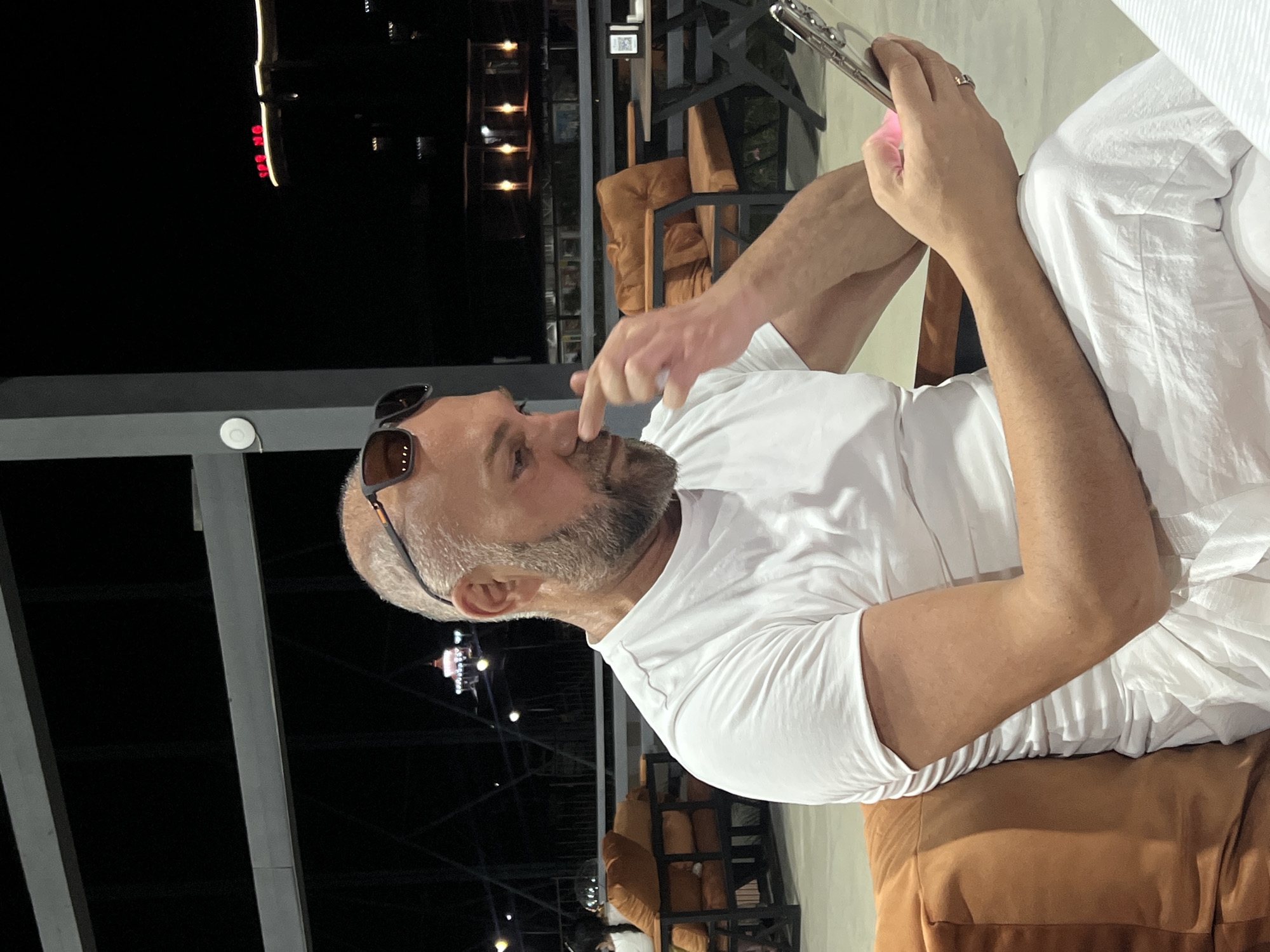 A bald, middle-aged man with a gray beard is seated on an orange cushion on an outdoor patio at night, wearing a white t-shirt and white pants. He is holding a smartphone in his right hand and touching his nose with his left index finger. The background shows outdoor seating with more orange cushions, tables, and in the distance, city lights and buildings, including one with red neon signage. The scene suggests a relaxed, possibly urban, evening setting. The image was captured in Sabunchu Raion, Azerbaijan.