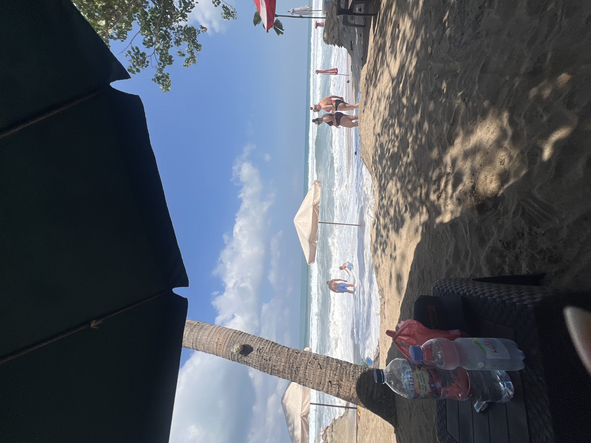 This is a ground-level, eye-level shot of Kuta Beach, Bali, Indonesia, on a bright, sunny day. The foreground is dominated by dark sand, with patterned shadows cast by palm trees and umbrellas. A black plastic table in the lower left corner holds two water bottles, one large and clear with a blue cap and the other smaller and frosted with a pink label, along with a red plastic bag and a black baseball cap.
In the mid-ground, a weathered palm tree stands to the left, its trunk textured and appearing to have some objects attached to it. The ocean is visible behind, with moderate waves crashing onto the shore. Two people are wading in the shallow water near the palm tree: an adult male wearing blue shorts and a young child.
Further back, on the sandy shore, two beach umbrellas stand, one light tan and the other white. To the right, two women in swimsuits are standing on the sand, facing away from the camera towards the ocean. In the background, the ocean stretches to the horizon under a blue sky with scattered white clouds. Two red and white umbrellas are visible further down the beach, along with some sandbags. The overall scene depicts a relaxed beach environment with people enjoying the water and the sun.