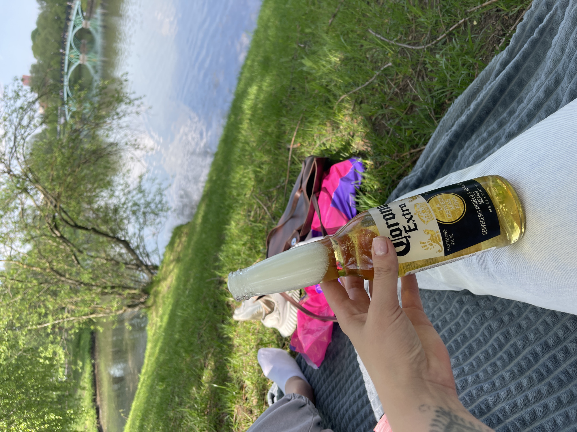 Here is a description of the image:
A person is seen relaxing by a river on a sunny day, holding an open bottle of Corona Extra beer. They are sitting on a gray textured blanket placed on grass. The person is wearing light gray pants and white socks with dark sneakers visible nearby. Next to them is a brown leather bag and a bright pink and purple umbrella. The bottle of beer is foaming over, held in the person's right hand, which has a tattoo on the wrist.
In the background is a river and lush green vegetation. A bridge with distinctive arch-like supports is seen in the distance.
Based on the architectural style of the bridge and the general environment, it is likely the location is Moscow, Russia.