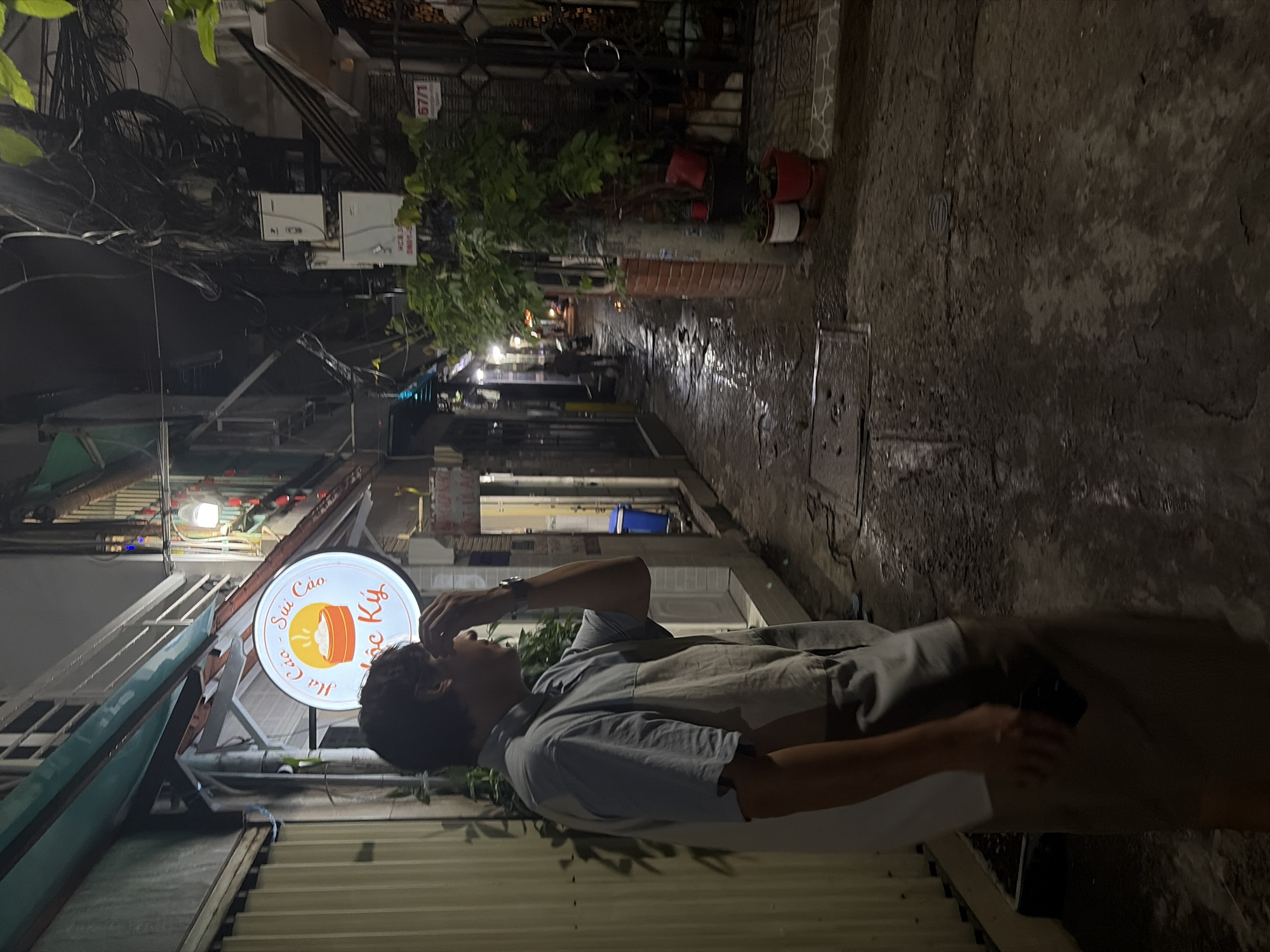 This image captures a nighttime scene within a narrow urban alleyway in Thủ Đức, Vietnam. The ground, primarily concrete, appears wet and uneven, reflecting ambient light, suggesting recent rainfall or cleaning.
In the foreground, slightly left of center, stands a male individual. He has short, dark hair and is wearing a two-toned, short-sleeved collared shirt, light blue on top and a lighter, possibly beige or grey, on the bottom, paired with light brown or khaki pants. A watch is visible on his left wrist. His left hand is raised to his forehead, partially obscuring his face as he looks towards the right side of the alley, away from the camera. His right hand holds a dark-colored smartphone, hanging loosely at his side.
To the immediate left of the man, a circular, illuminated sign for a food establishment named "Há Cảo - Sủi Cảo Ngọc Ký" is affixed to a building. The sign features a graphic of a steaming bowl of dumplings. The building on the left has a section with corrugated metal siding.
On the right side of the alley, buildings feature a dark, ornate metal gate or door. A white sign with the number "67/1" is attached to the building structure, identifying the address. In front of this building, several potted plants, including red and white planters, are arranged near a brick-patterned base. Overhead, a dense network of electrical and communication cables is visible, tangled and interconnected, with several utility boxes mounted on a pole or wall structure.
The alley extends into the background, where more ambient lights are visible, indicating further establishments or residences. A blue trash bin is discernible further down the alley on the right side. The overall environment suggests a bustling, densely populated urban area, characteristic of Vietnamese cities.