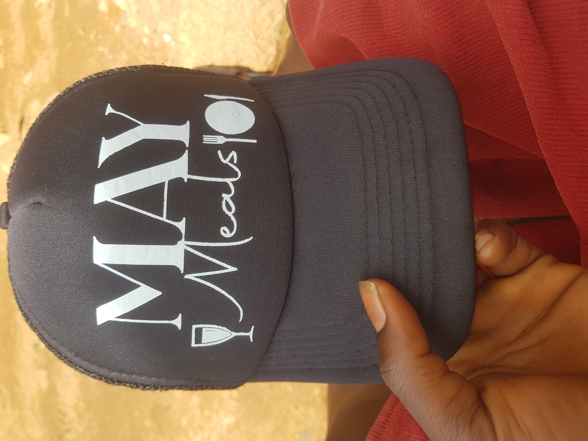 A close-up, overhead shot shows a person's hand holding a black trucker hat. The hat has a white graphic on the front that reads "MAY Meals" with icons of a wine glass, a fork, and a plate. The background is blurred, but appears to be a sandy, outdoor surface, possibly in a city. The person's hand is dark-skinned and is holding the hat from the side. The hat is made of a mesh material in the back and a solid fabric in the front. The overall lighting suggests it is daytime.