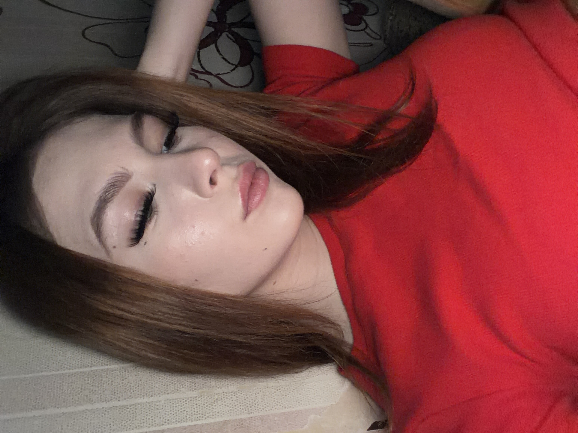 A young woman with light skin, long brown hair, and well-defined eyebrows and eyelashes is lying on her back. She is wearing a red top. The background appears to be a patterned surface, possibly bedding. The location is not discernible as Noviy, Russia from the given image.