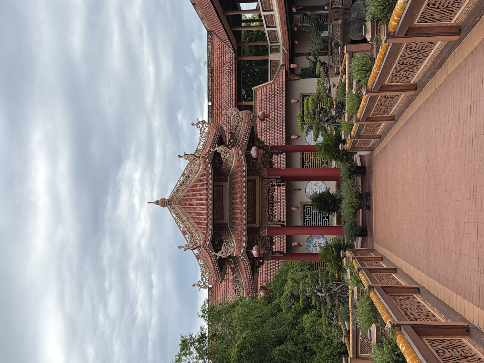 A red brick pathway leads towards a traditional East Asian building with a multi-tiered, red-tiled roof and ornate detailing. The building is supported by thick red pillars and features red lanterns hanging from its eaves. Lush green trees and manicured bonsai plants surround the structure. The sky above is overcast with gray clouds. This image was taken at the Quan Am Pagoda in Hoi An, Vietnam.