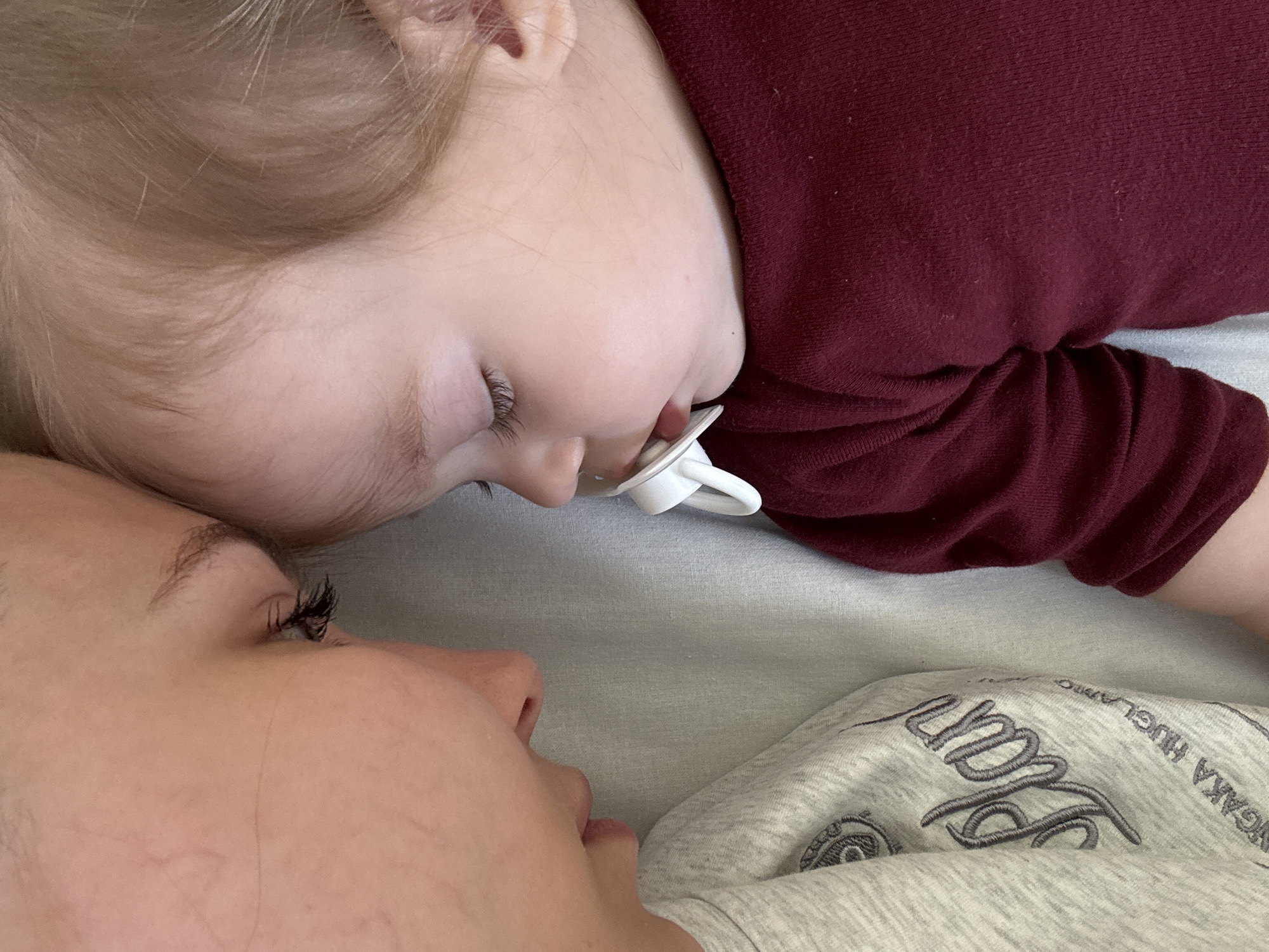 The image displays a close-up, overhead view of an adult and an infant or young child in a shared, intimate moment. The precise geographical location, including any specific city such as Chaikovsky in Russia, cannot be determined from the visual information provided.
On the left, an adult, presenting as female, is depicted in profile, lying on their side with eyes closed or downcast. They have fair skin, dark, prominent eyelashes, and are wearing a light grey long-sleeved top. Partial text embroidery on the shirt, including the word "Plan" and "HUGGLABBO," is visible on the left side.
On the right, an infant or young child is also depicted in profile, lying on its side with eyes closed. The child has fair skin, light blonde hair, and is wearing a plain maroon long-sleeved top. A white pacifier is held in its mouth.
The adult's forehead is very close to or lightly touching the child's forehead, suggesting an embrace or shared moment of rest. Both individuals are positioned on a light grey or off-white fabric surface, likely bedding, which appears soft and slightly rumpled. The lighting is soft and diffused, indicating an indoor setting.