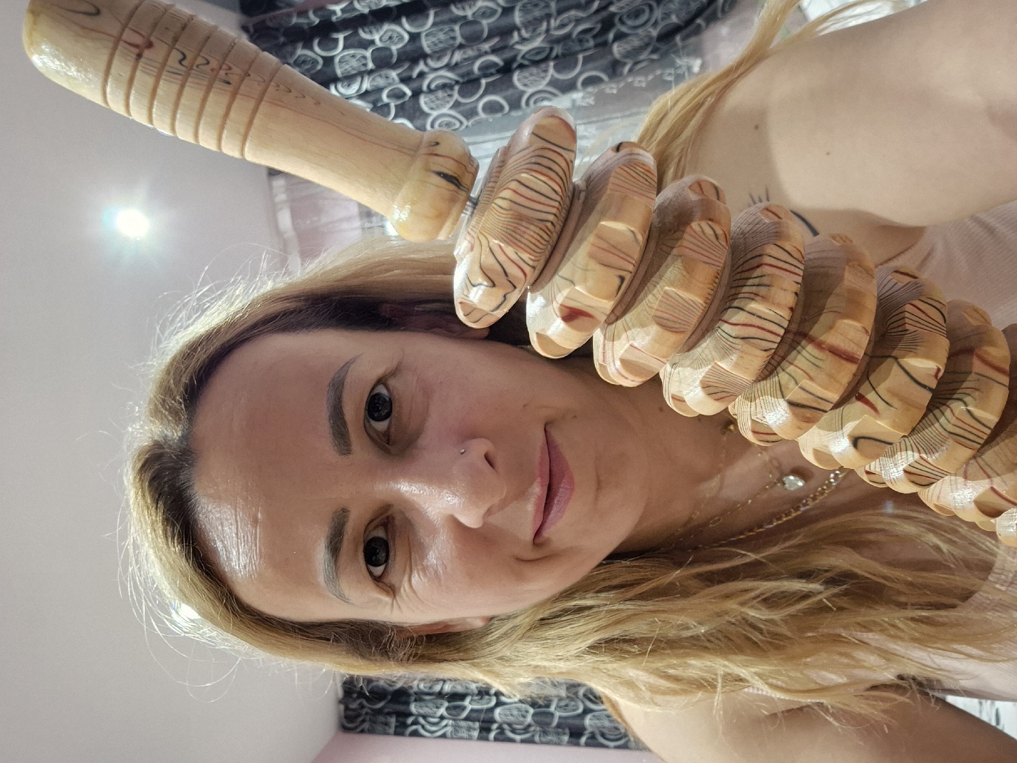 A selfie of a woman holding a wooden massage tool in front of her face. The tool has a handle and a series of ribbed rollers. The woman has blonde hair, fair skin, and is wearing a necklace. The background is blurred, showing a patterned curtain and a light-colored wall. Location: Unknown, Unknown.