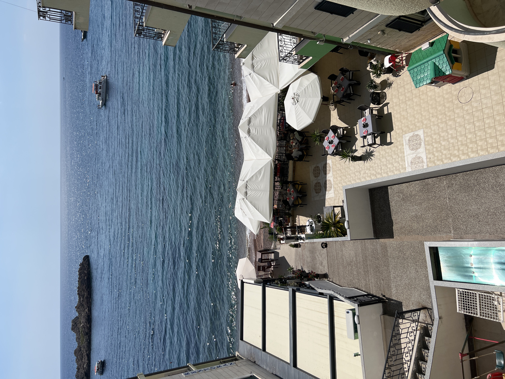 The image provides an elevated view of a coastal area in Sveti Stefan, Montenegro, featuring a restaurant with outdoor seating and the adjacent sea. The scene is illuminated by clear sunlight.
In the foreground, to the left, are several levels of building structures with awnings. Below, a stairway with a decorative railing leads down. Further down, a section of a turquoise water feature is visible, along with a child's swing set and a metal grate.
The central area of the image showcases a courtyard and dining area. Multiple tables with chairs are arranged under large white umbrellas. Some tables are set with plates and red napkins. Several potted plants are placed around the seating area. A small, colorful playhouse is situated on the tiled ground to the right.
The background depicts the blue sea, with a gentle shoreline. A few boats are visible on the water, one larger vessel with people on its deck, and a smaller one near a rocky island formation. In the sea, a single person is swimming near the shore. The horizon line is clear with a bright sky above. The overall atmosphere is one of a sunny, leisurely day at a seaside establishment.