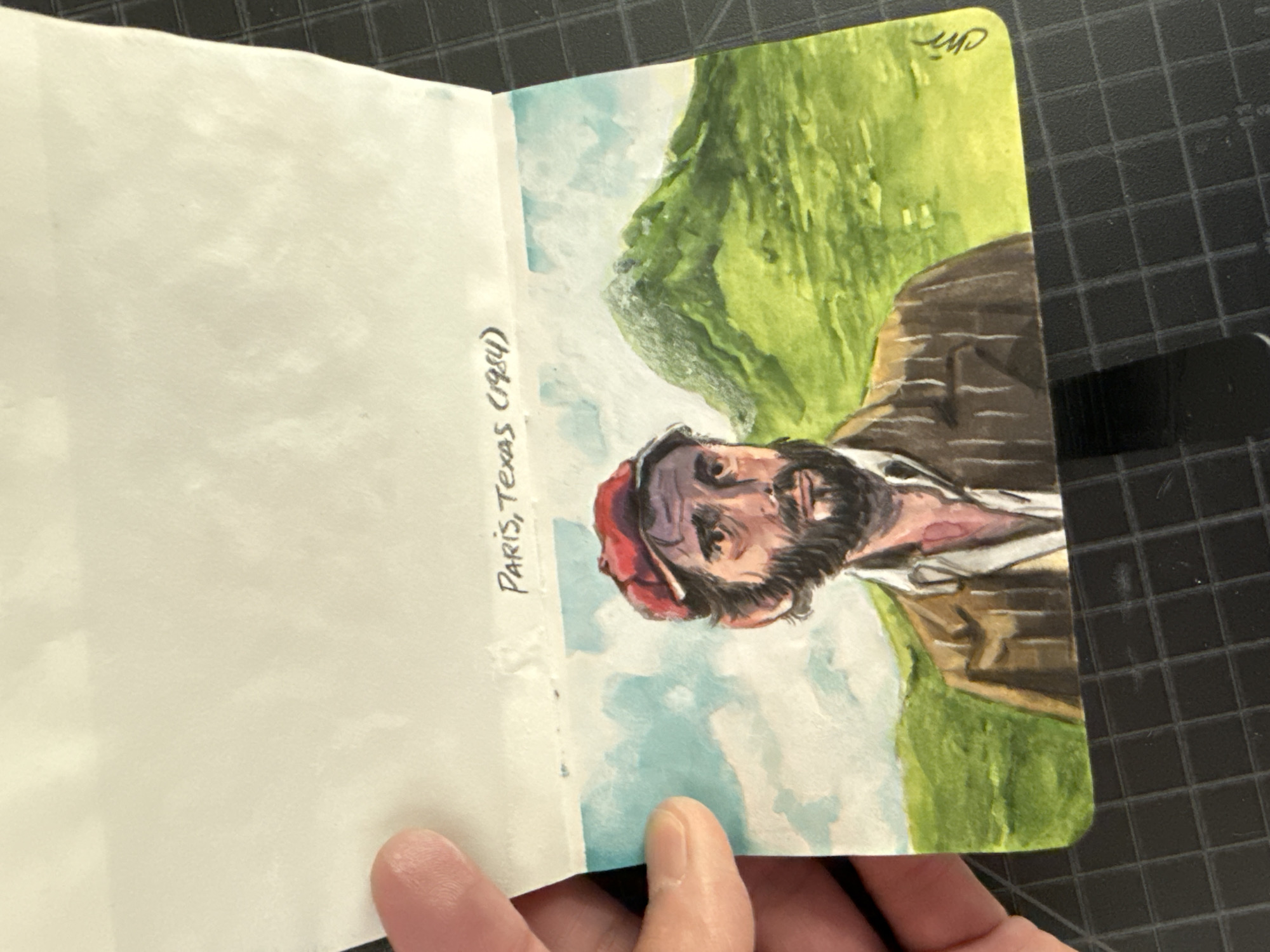 The image shows an open sketchbook. The left page is blank. The right page contains a watercolor painting of a bearded man wearing a red hat and a striped jacket, set against a backdrop of green hills and a light blue sky. The text "Paris, Texas (1984)" is written in black ink on the left page, adjacent to the painting. The sketchbook is resting on a black gridded surface. A person's hand is holding the bottom of the sketchbook. The location is irrelevant, as it is not visible in the picture.
