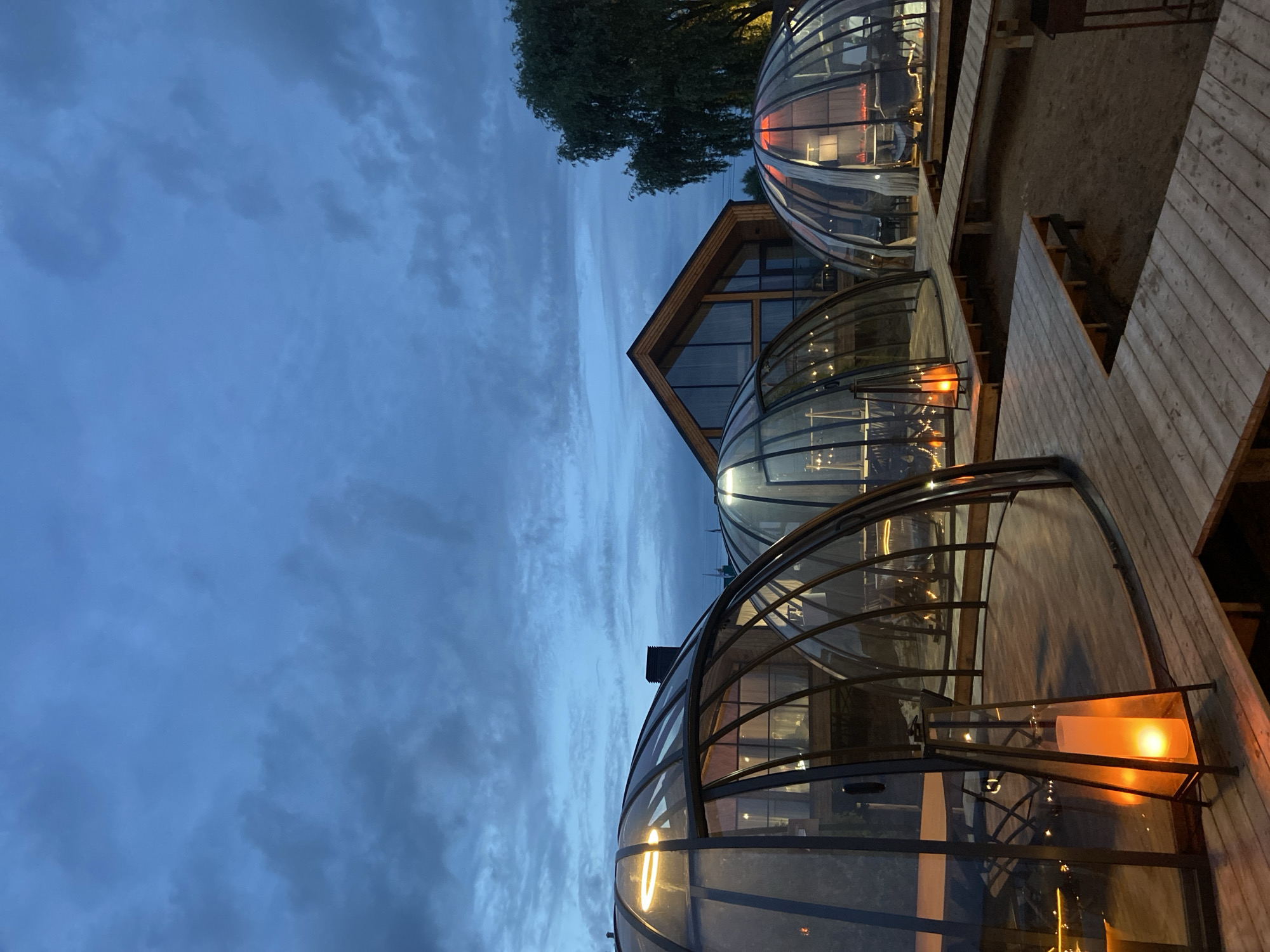 The image depicts a row of glass-enclosed dining pods, possibly igloos or orangeries, situated on a wooden deck at dusk. The sky is a deep blue with dramatic clouds, suggesting twilight or an impending weather change. The pods are illuminated from within, casting a warm, inviting glow that contrasts with the darkening sky. Inside the pods, tables and chairs are visible, along with decorative lights and what appears to be a candle in a lantern in the foreground pod, creating a cozy and intimate atmosphere.
Behind the pods, a wooden structure with large windows and a pitched roof is partially visible, hinting at a larger building or restaurant. A large tree with dark green foliage stands in the background, adding to the natural setting. The wooden deck has a slightly weathered appearance and some areas are covered in sand or fine gravel, suggesting an outdoor or semi-outdoor dining area. Stairs are also visible leading down from the deck.
The scene evokes a sense of calm and relaxation, with the lighting and the enclosed nature of the pods suggesting a comfortable and perhaps romantic dining experience. The time of day is clearly evening, and the weather, while cloudy, does not appear to be actively raining. There is no visible text in the image. The location context of Votkinsk, Russia, suggests a cool climate where such enclosed dining structures might be popular during certain seasons.