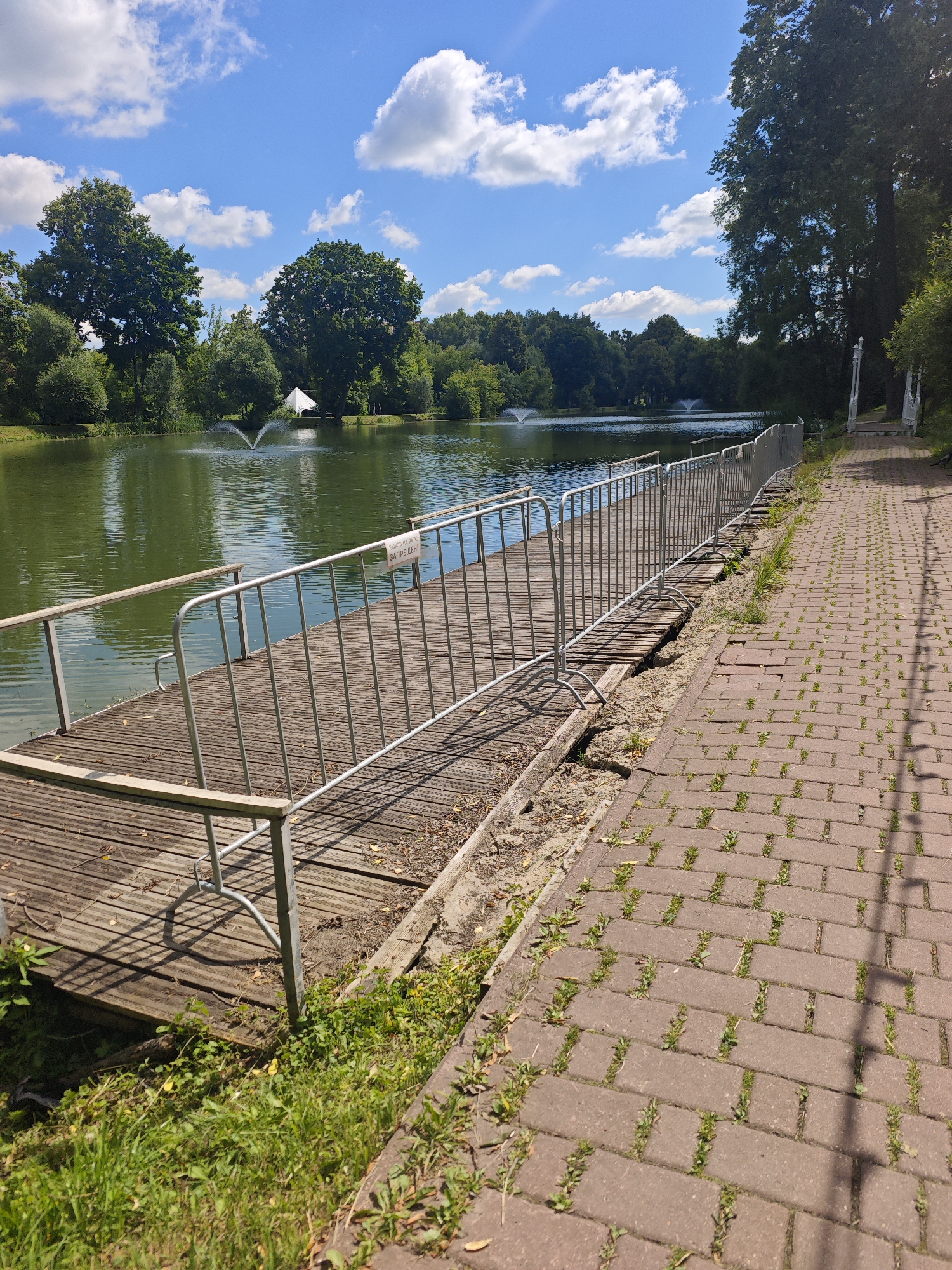 The image depicts a wooden pier extending into a calm, green body of water, likely a lake or pond, on a bright, sunny day. The water features two fountains spraying upwards. A paved pathway runs alongside the pier, bordered by grass and trees in the background. The pier is equipped with a metal railing for safety, and a sign is attached to it, though the text is illegible. A white tent or structure is visible in the distance on the opposite bank. The scene appears to be in a park or recreational area, suggesting it could be within the city of Chekhov, Russia, given the context of the prompt. There are no people visible in the image.
