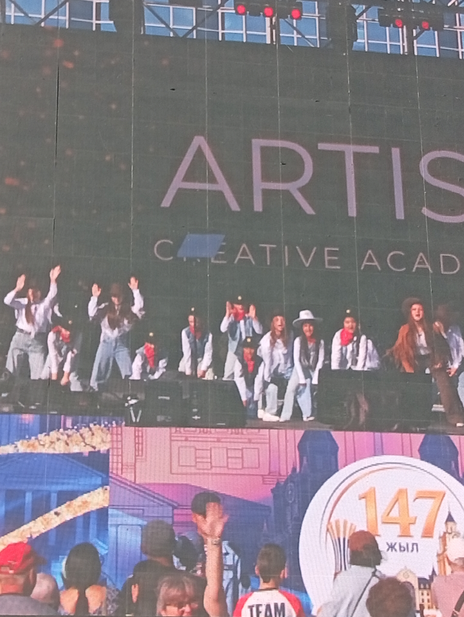 The image displays a large LED screen at an outdoor event. On the screen, the words "ARTIS CREATIVE ACAD" are visible, followed by a circular emblem containing the number "147" and the word "ЖЫЛ" (which means "year" in Kazakh). Below the screen, a crowd of people are gathered, facing the screen. Some individuals in the crowd are wearing hats, and one person in the foreground is wearing a red cap. The overall impression is that of a performance or ceremony taking place at an open-air venue, possibly a concert or festival. The event is likely in Kostanay, Kazakhstan, given the inclusion of the Kazakh word "ЖЫЛ".