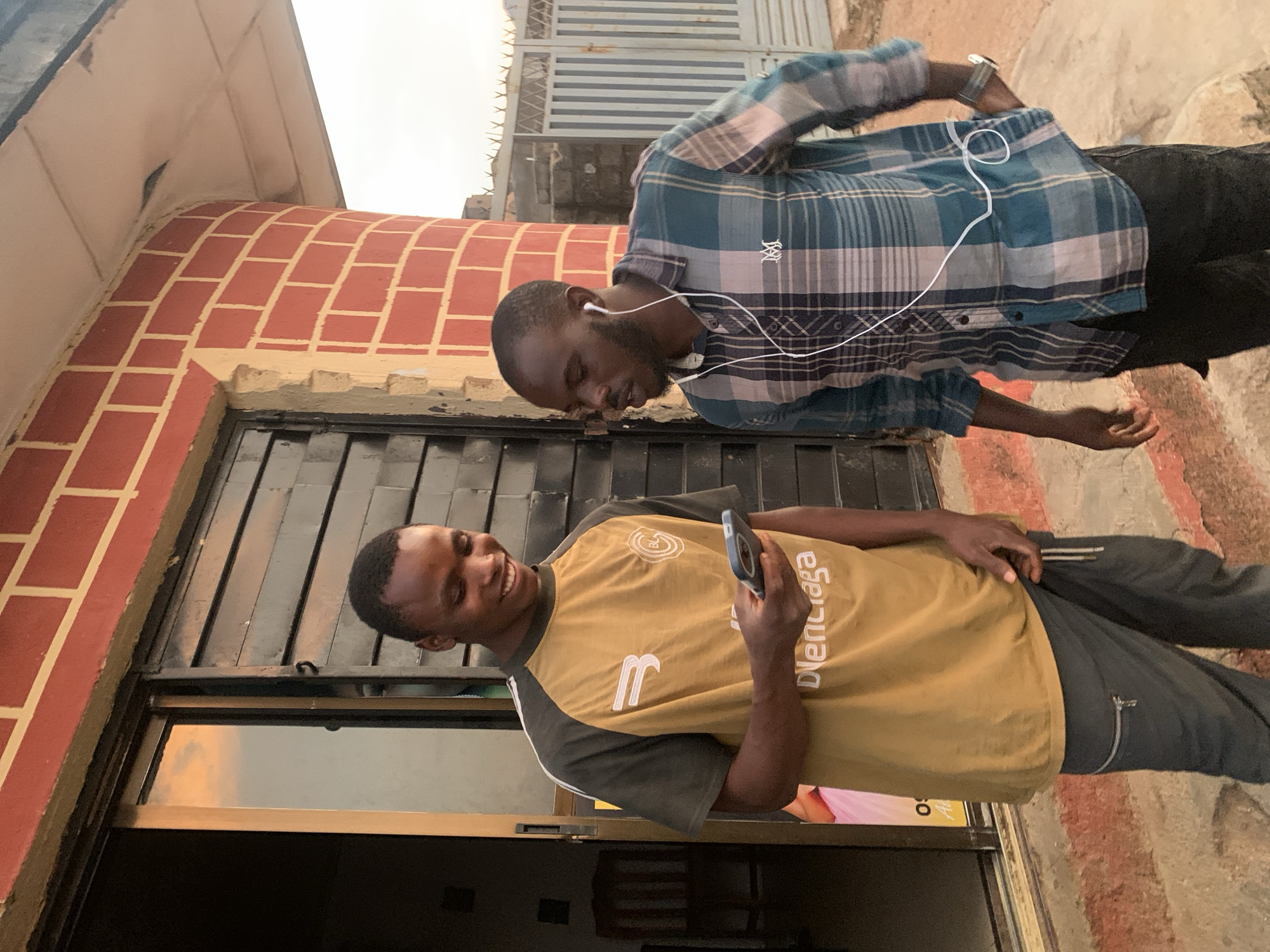 Two young Black men stand outside a building in Jos, Nigeria. The man on the left, wearing a mustard-yellow shirt with dark gray sleeves and gray pants, smiles as he looks at his phone. The man on the right, dressed in a blue and white plaid shirt and black pants, wears white earphones and appears to be speaking. The building behind them is constructed with red bricks and has a dark metal door. A white fence and more buildings are visible in the background. The ground is a rough, reddish-brown surface. The lighting suggests it is late afternoon or early evening.