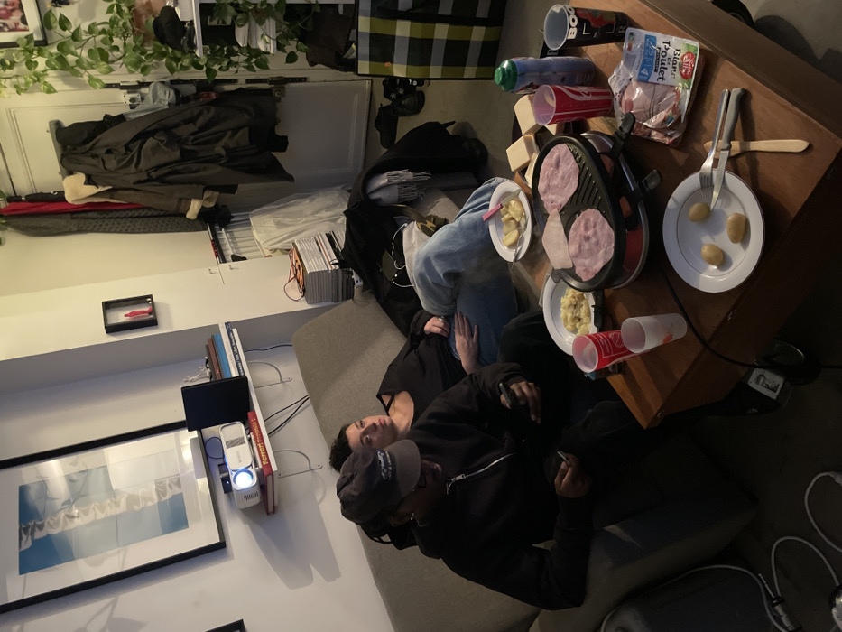 Two people are lounging on a gray couch. A man wearing a black jacket and baseball cap sits up holding a phone, while a person in jeans and a black top reclines. A wooden table is laden with food: sliced ham, potatoes, plates with potatoes and cheese, disposable cups, and a small raclette grill. The room includes a framed picture, projector, and a shelf of books. A plant hangs near a shelf with clothes.