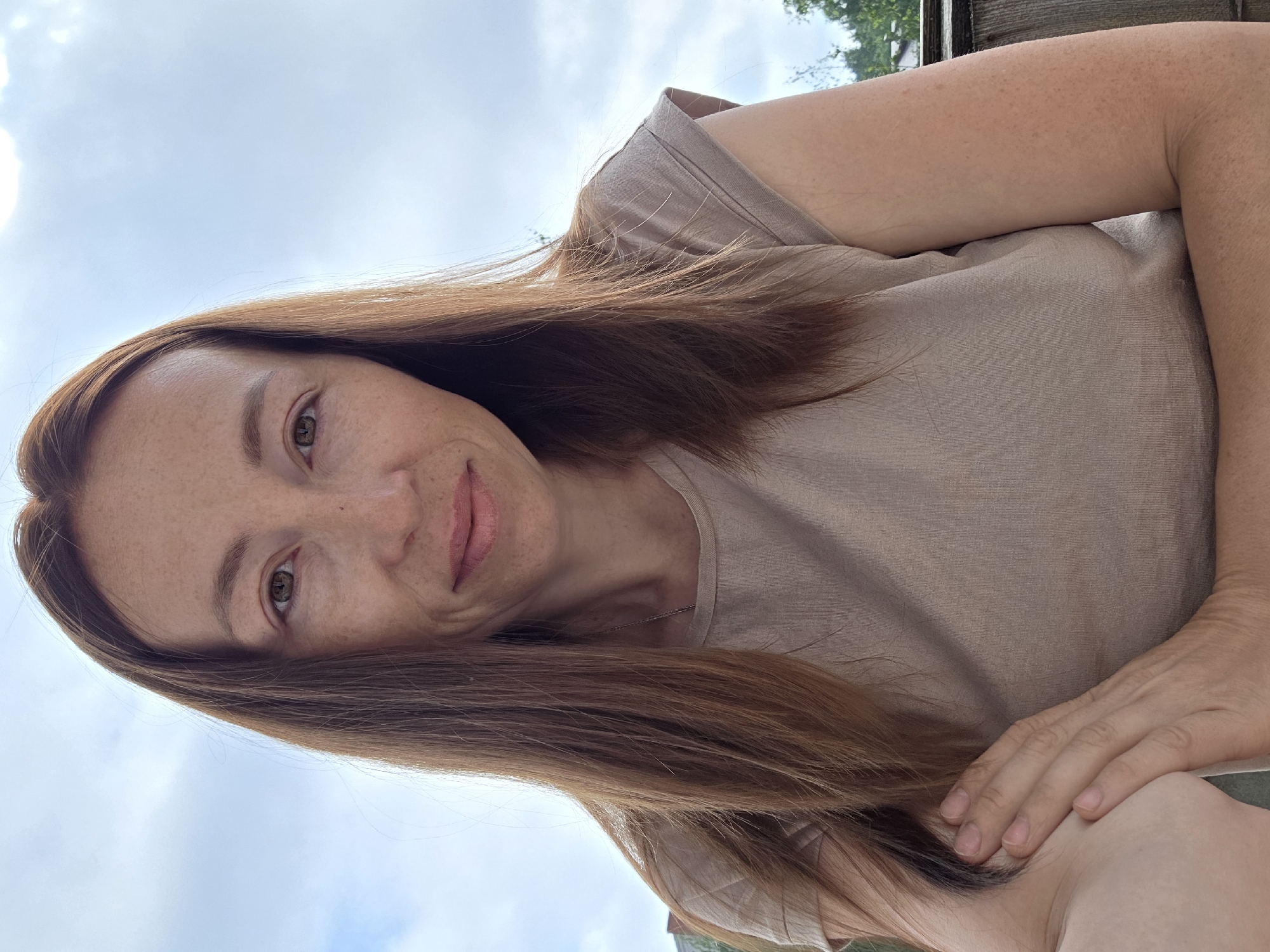 The image shows a close-up, waist-up portrait of a woman with long, auburn hair, freckles, and light brown eyes. She is wearing a light brown t-shirt and has her arms crossed. The background consists of a cloudy sky and some greenery, suggesting an outdoor setting. The image was taken in Chaikovsky, Russia.