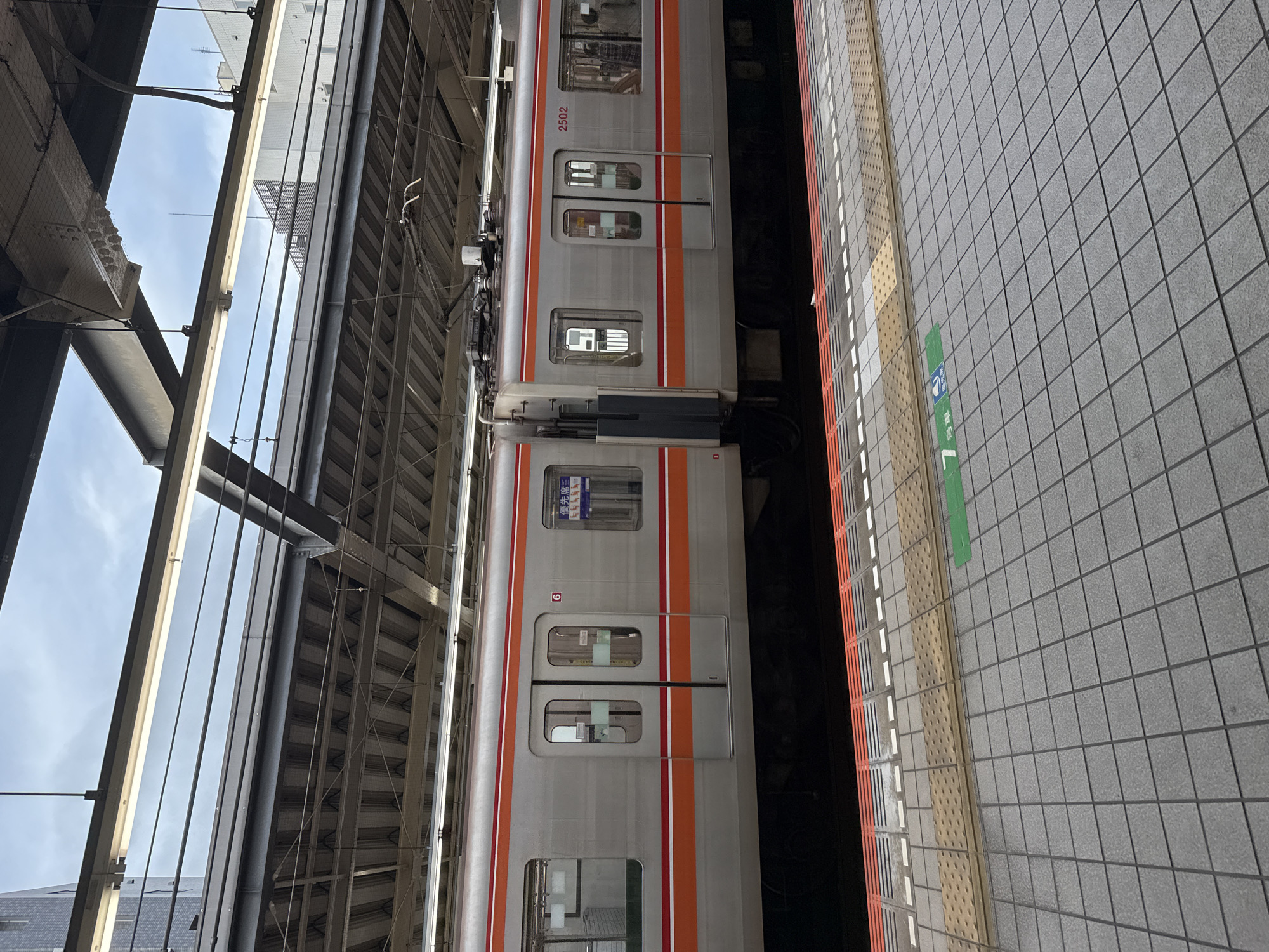 An exterior medium shot captures a silver train with orange and white stripes at a station platform in Yachiyo, Japan. The train is stationary, with its doors closed. The platform is paved with grey tiles and features tactile paving for visually impaired individuals. A green sign indicating "7 car" and "priority seat" is visible on the platform. The station appears to be covered by a modern, multi-layered roof structure with exposed beams and lighting fixtures. The sky above is overcast. The scene is captured from a slightly elevated, eye-level perspective, focusing on the train and platform.