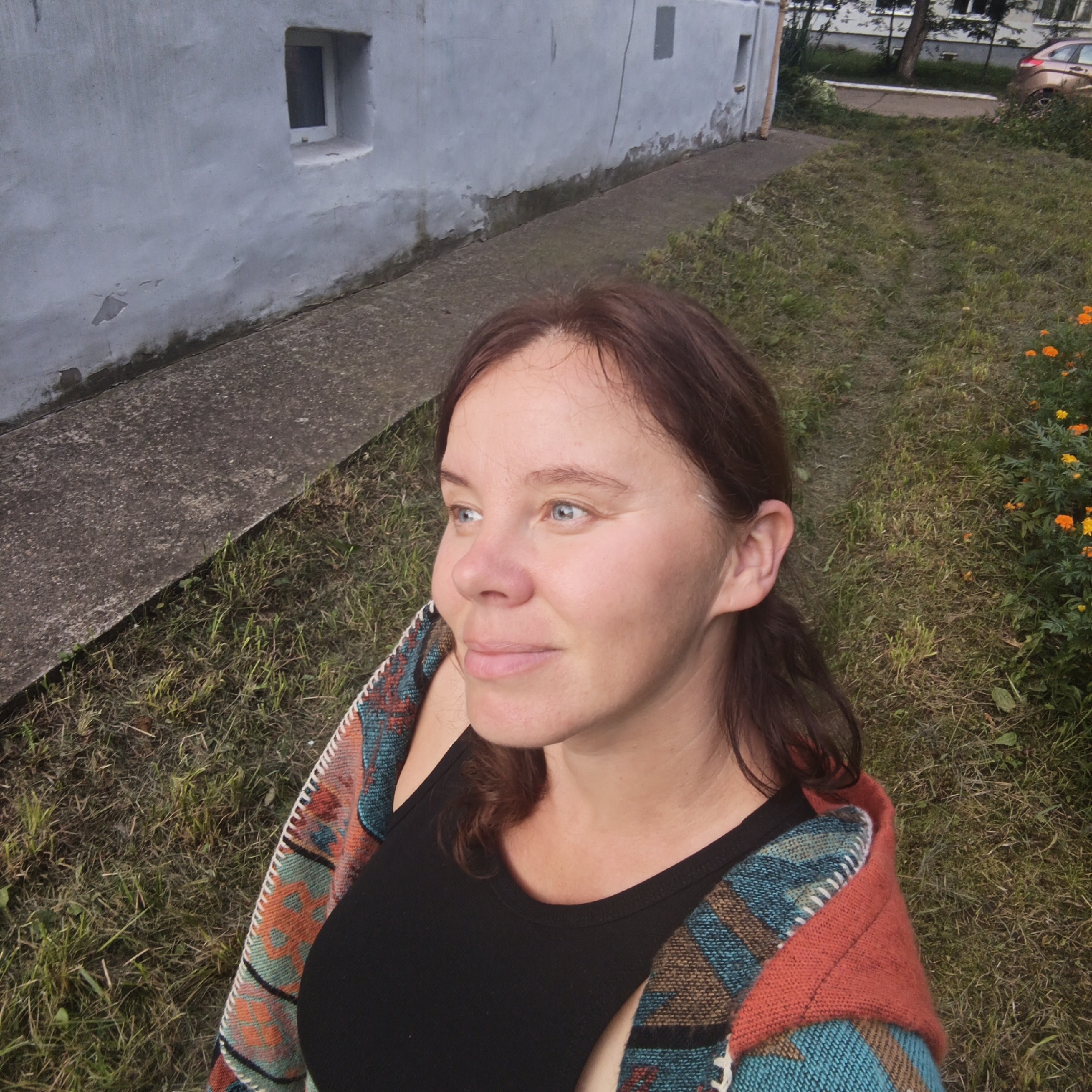A medium-shot portrait of a woman with light skin, brown hair, and blue eyes is captured outdoors, looking to her left. She is wearing a black tank top and a colorful, patterned blanket draped over her shoulders. The background consists of a light-colored building with a small window and a concrete walkway, bordering a grassy area with scattered orange and yellow flowers. The location is identified as Chaikovsky, Russia.