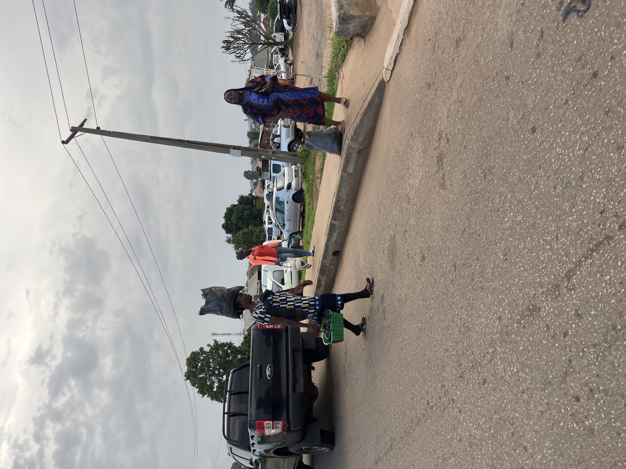In Bukuru, Nigeria, a woman walks down a paved road carrying a large bag of charcoal on her head and a green basket of charcoal in her hand. She is wearing a patterned dress and black sandals. To her right, a woman in a blue and red patterned dress stands on a sidewalk next to a black trash bag and a utility pole. Several cars are parked on the right side of the road. A black pickup truck is parked on the left side of the road, and a portion of its rear is visible. A second woman in an orange jacket and blue jeans walks away from the camera on the sidewalk in the distance, carrying a white bucket. The sky is overcast with gray clouds.