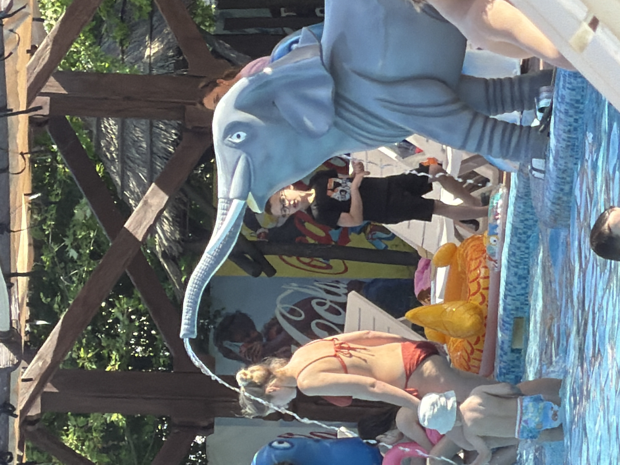 The image captures a poolside scene on a sunny day in Sociteni, Moldova. In the foreground, a woman in a red bikini is partially submerged in the blue tiled swimming pool, with water splashing from an elephant-shaped fountain. To her left, a young child wearing a white cap and patterned swim trunks is also in the water. Behind them, several inflatable pool toys, including a yellow duck and an orange fish, are visible.
The background reveals a wooden structure, possibly a canopy or a part of a poolside bar, with lush green foliage peeking through the gaps. Further back, a wall mural featuring a Coca-Cola logo and silhouetted figures adds a splash of color to the scene. A person wearing a black t-shirt is visible near the mural, partially obscured by the elephant fountain. The overall atmosphere appears relaxed and enjoyable, typical of a summer day at a swimming pool. The water is clear and reflects the bright sunlight, suggesting fair weather.